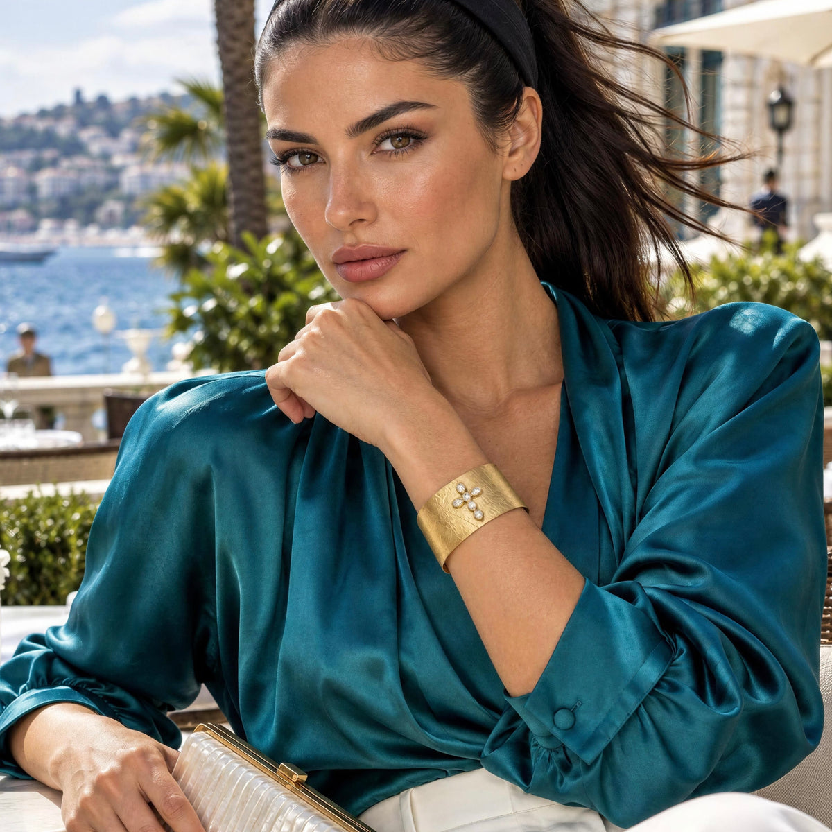 A woman wearing the Serafina Cuff from Chica Chica Jewelry and a shiny teal satin blouse, posed outdoors with a scenic coastal background.