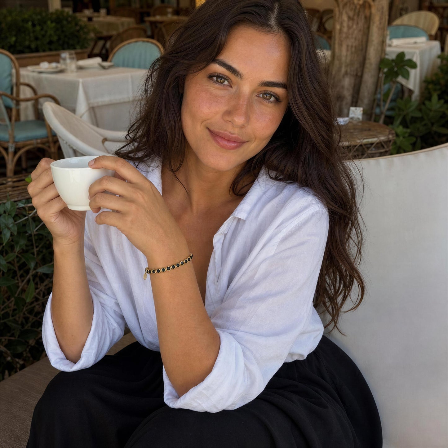 A woman with long dark brown hair sits at an outdoor cafe, holding a white ceramic cup. She is wearing a white button-down shirt, black pants, and the Siena Black Tennis Bracelet in her wrist.