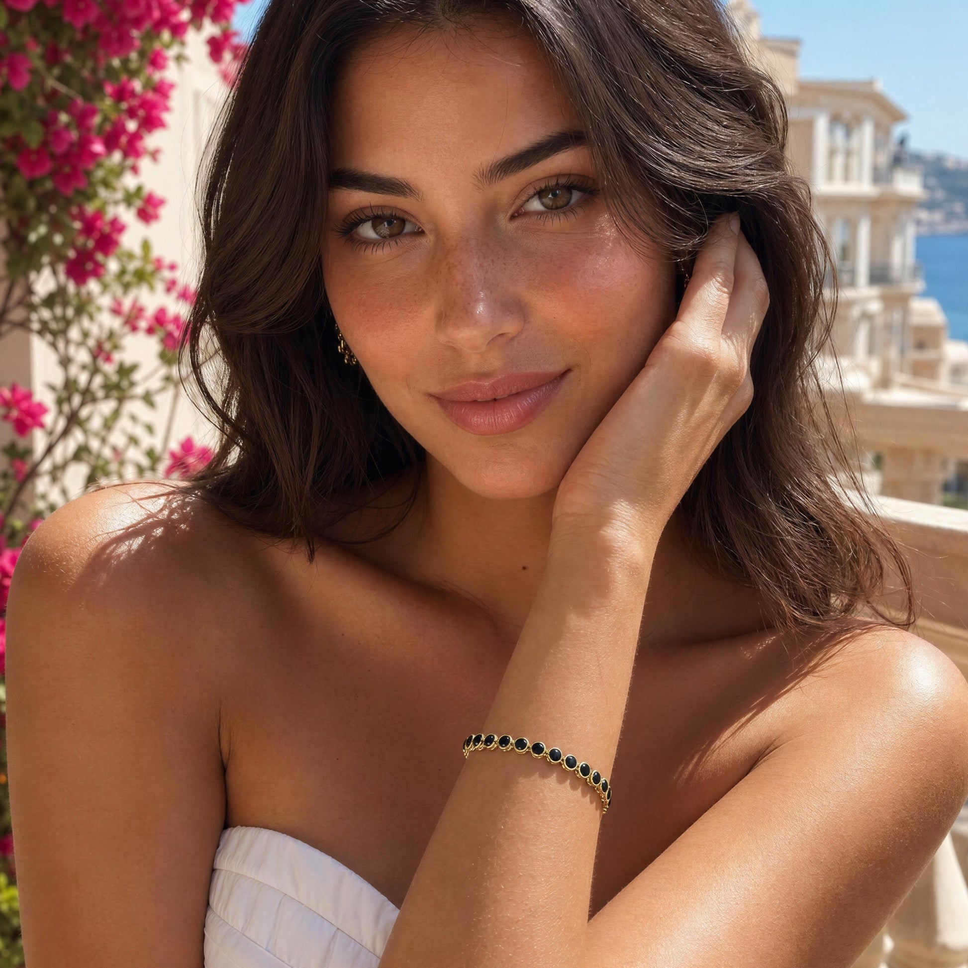 A woman wearing the Siena Black Tennis Bracelet posing outdoors with a white strapless top, pink flowers, and a coastal background.