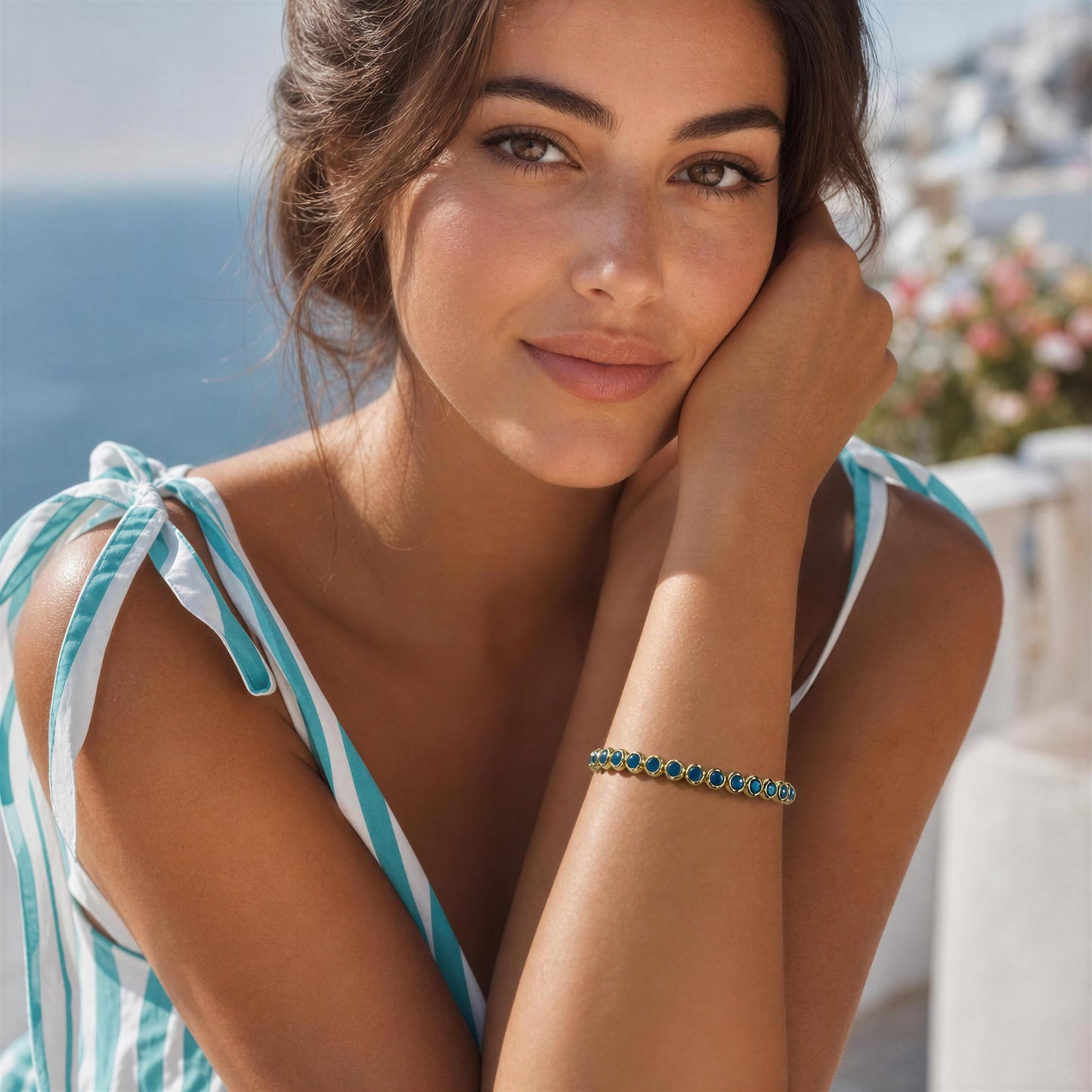 A woman outdoors wearing a delicate bracelet with small turquoise-blue stones set in a gold-tone band. She is also wearing a matching turquoise and white striped top, with a blurred coastal background.