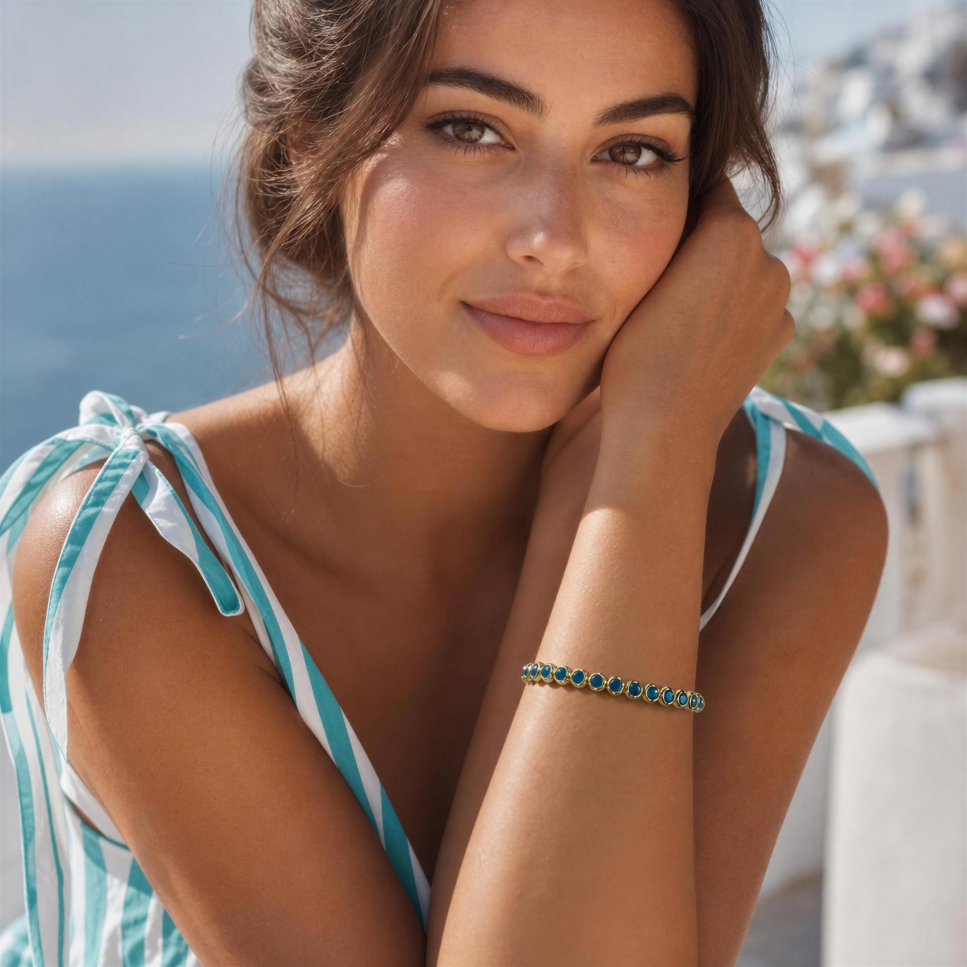 A woman outdoors wearing a delicate bracelet with small turquoise-blue stones set in a gold-tone band. She is also wearing a matching turquoise and white striped top, with a blurred coastal background.
