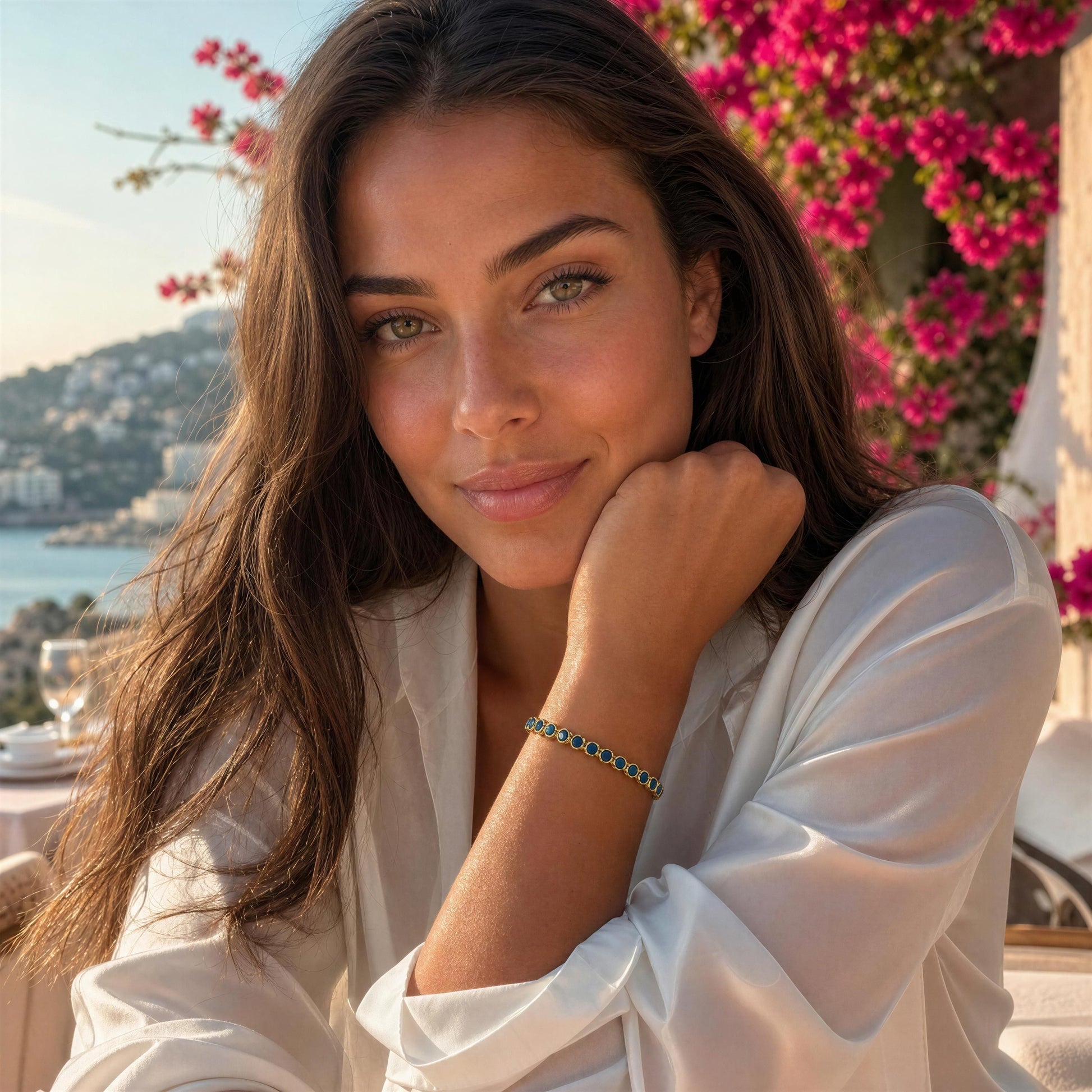 A woman with long brown hair wearing a white satin blouse and the Siena Turquoise Tennis Bracelet, posing outdoors with a scenic coastal background and pink flowers.