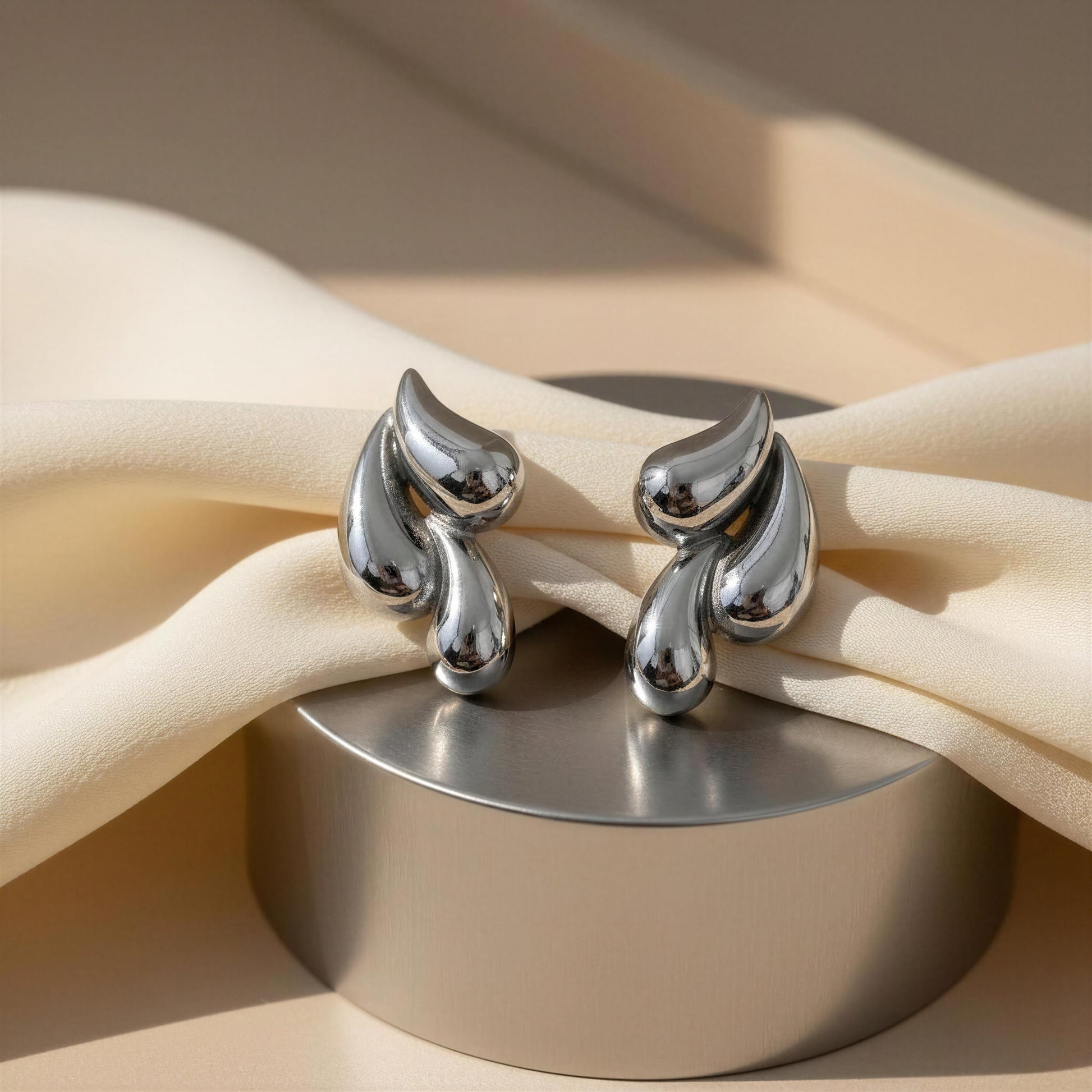 The Silver Molten Wave Drop Earrings on a beige fabric background