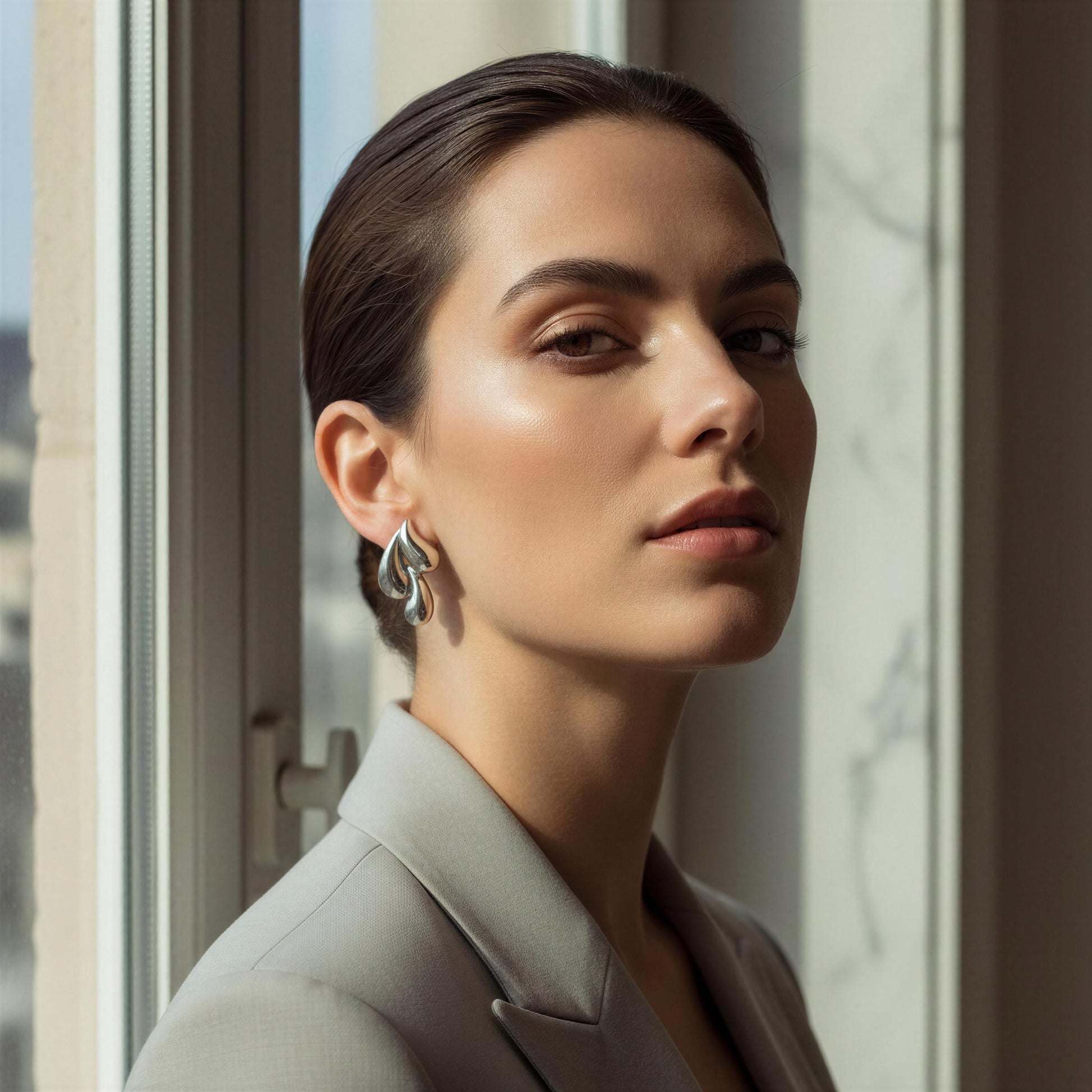 Woman wearing the Silver Molten Wave Drop Earrings standing by a window with a blurred cityscape background
