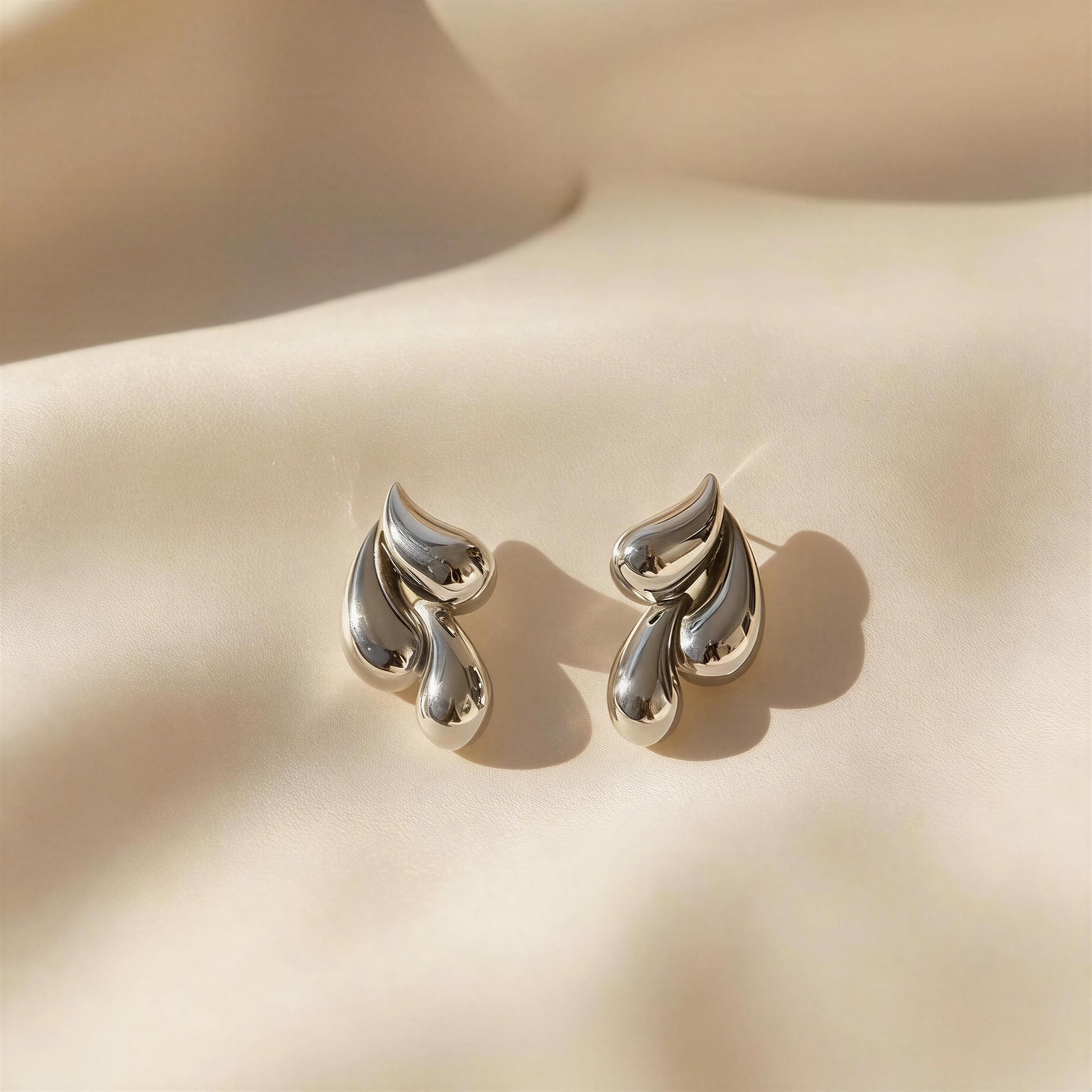 The Silver Molten Wave Drop Earrings on a beige fabric background