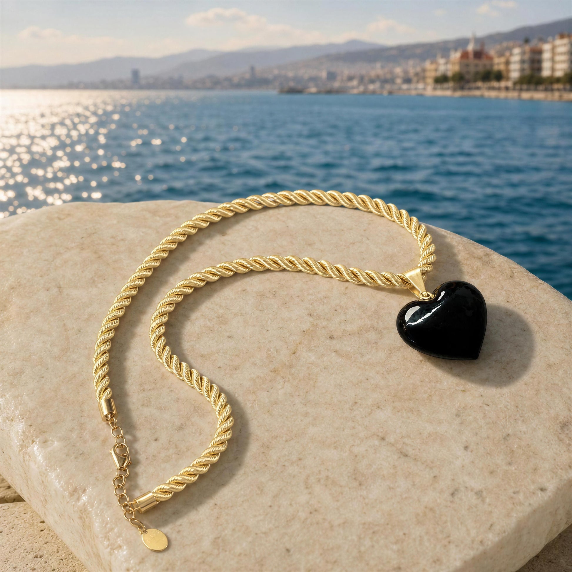 Close-up of the Siyah Murano-style black heart necklace resting on a natural stone by the Mediterranean coast, showcasing the twisted gold-tone cord, glossy black heart pendant, and adjustable extender.