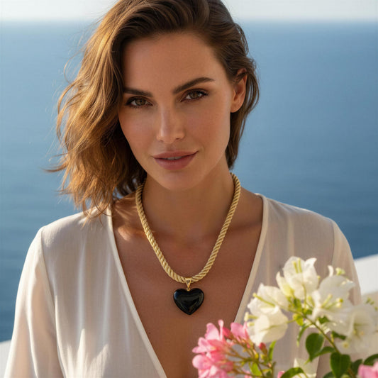 Woman wearing the Siyah Necklace featuring a black Murano glass heart pendant on a twisted gold-tone cord overlooking the Mediterranean coast.
