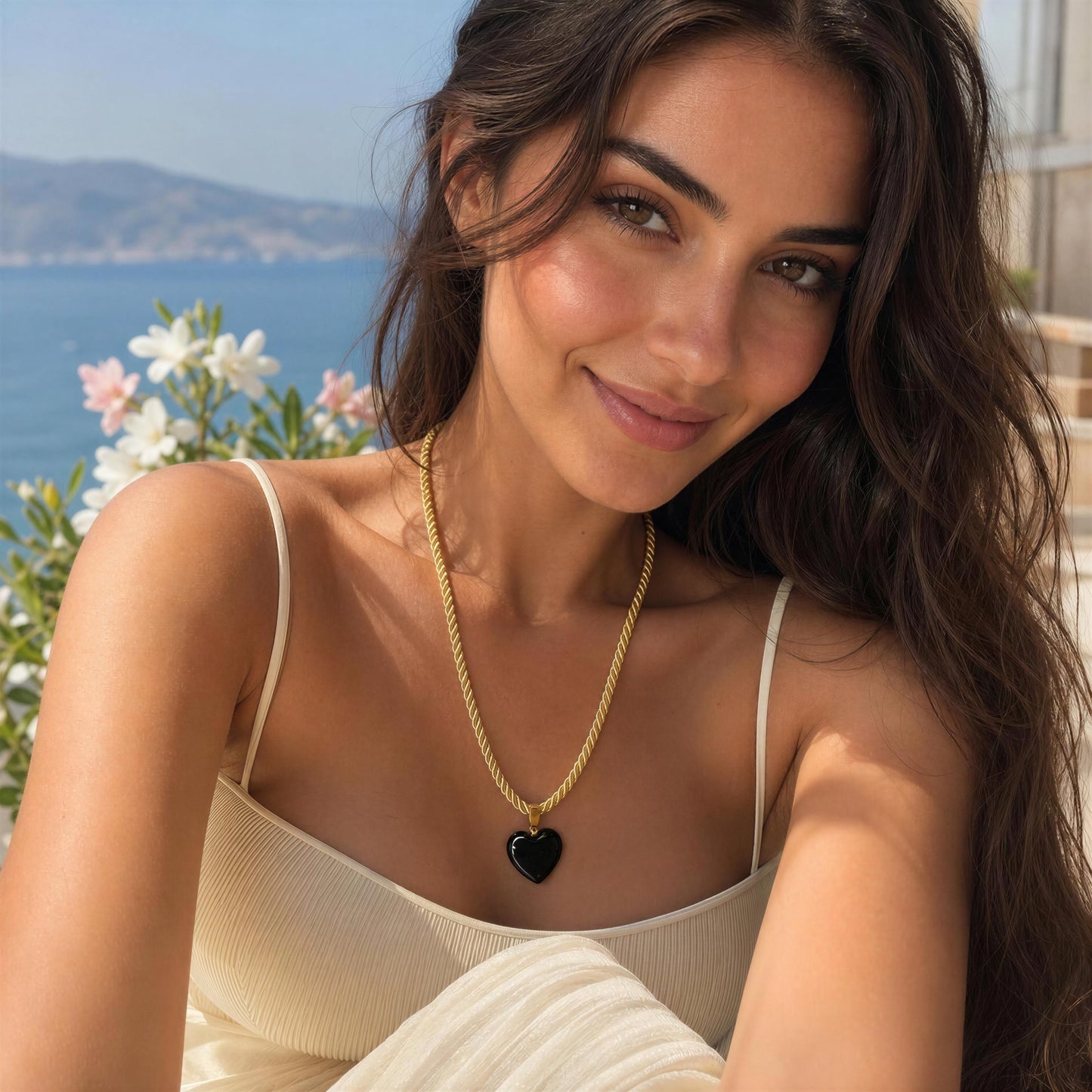 Woman wearing the Siyah Necklace featuring a black Murano glass heart pendant on a twisted gold-tone cord overlooking the Mediterranean coast.