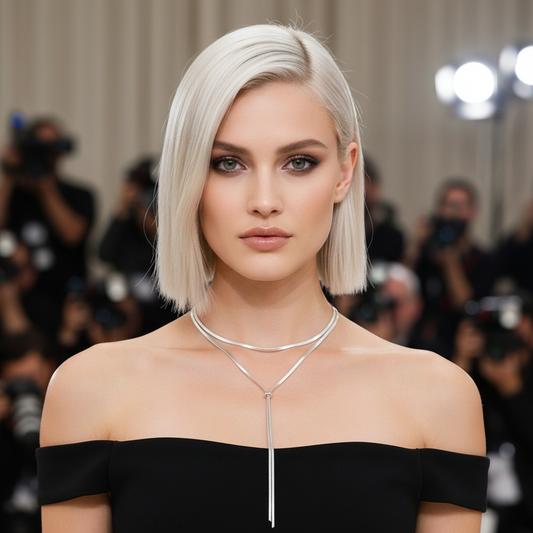 Woman with short blonde hair wearing a black off-shoulder dress and silver necklace in a blurred event setting.