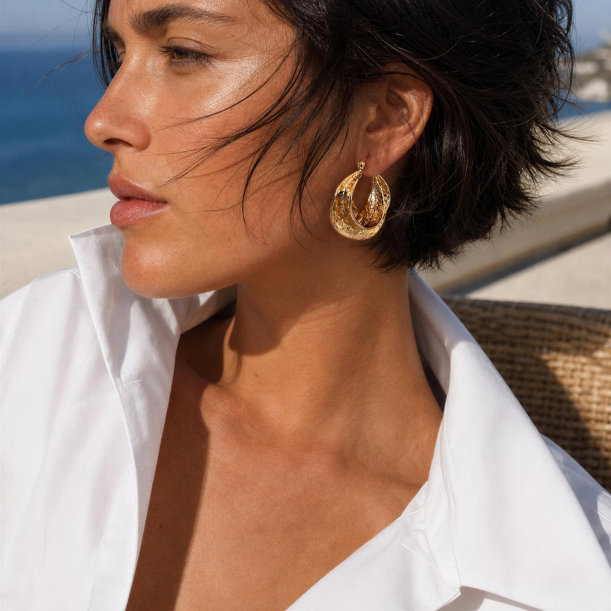 A close-up lifestyle shot of a woman with short dark hair wearing a textured gold hoop earring, a white collared shirt, and a white bikini top, with a beach and ocean background.