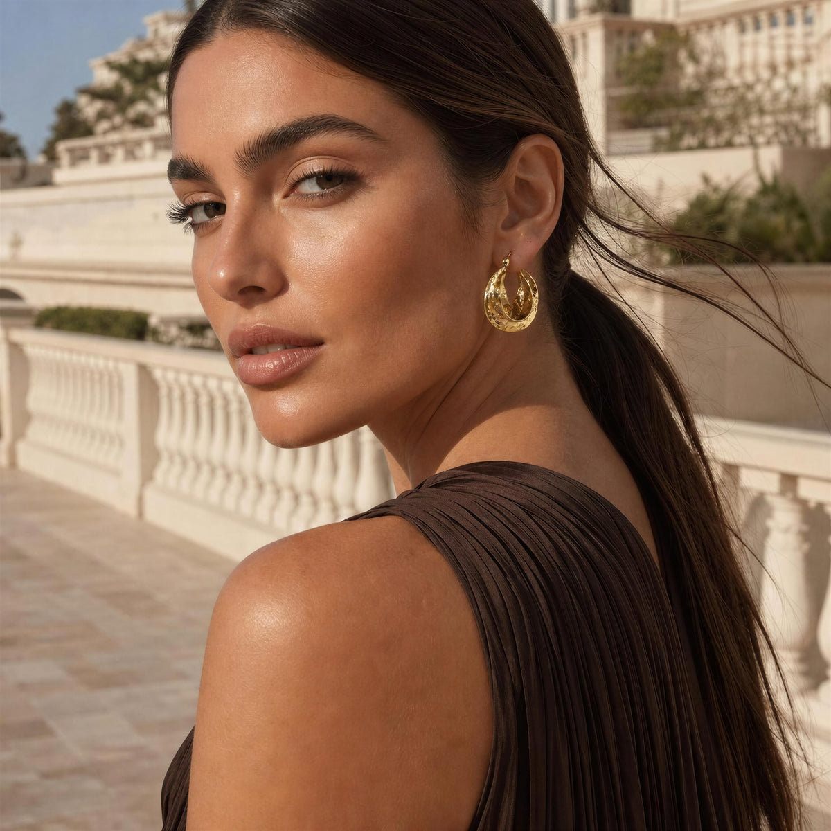 A woman wearing a textured gold crescent-shaped hoop earring, photographed from behind over her shoulder in an outdoor setting with a white balustrade and building in the background.