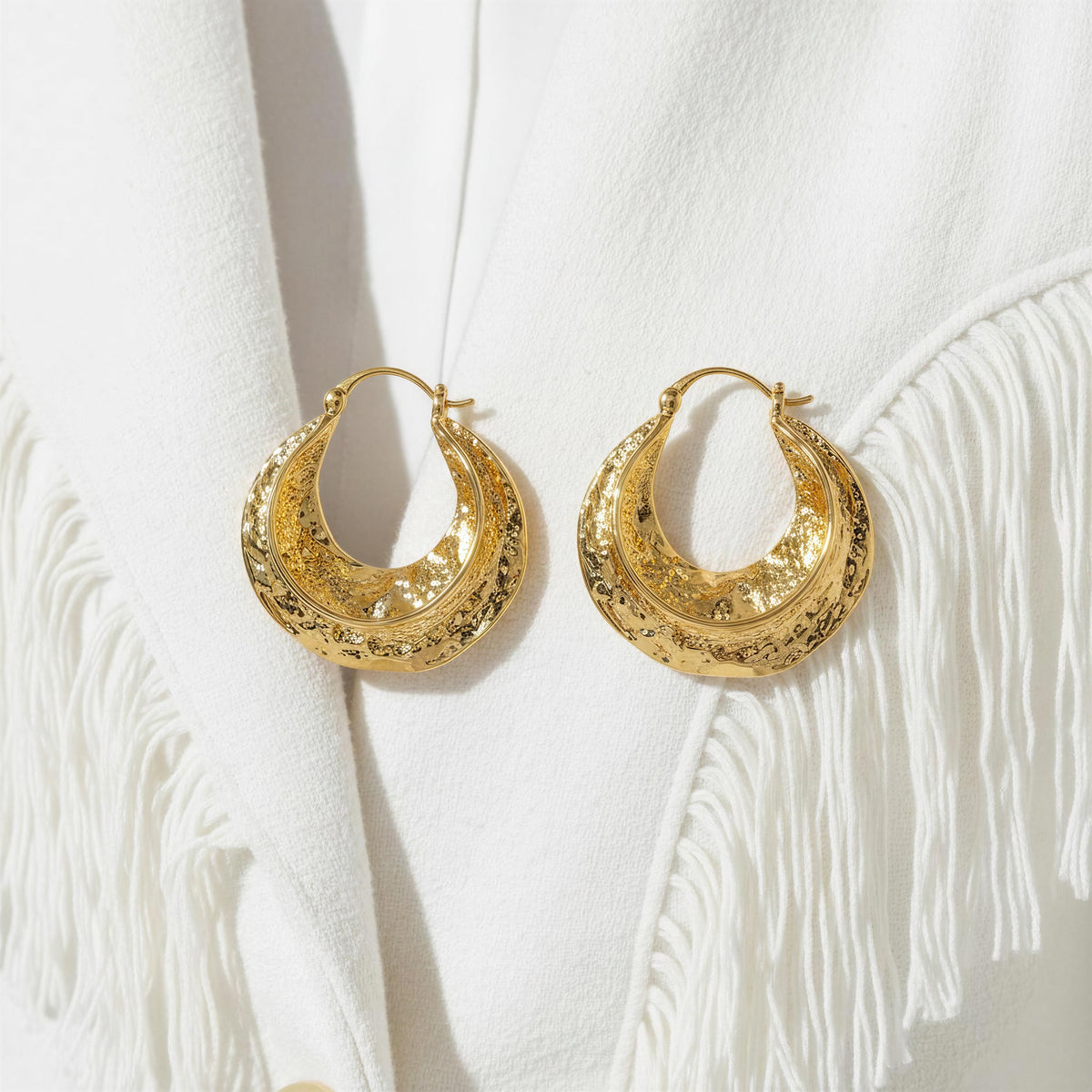 A pair of textured gold crescent-shaped hoop earrings displayed on a white fringed garment.