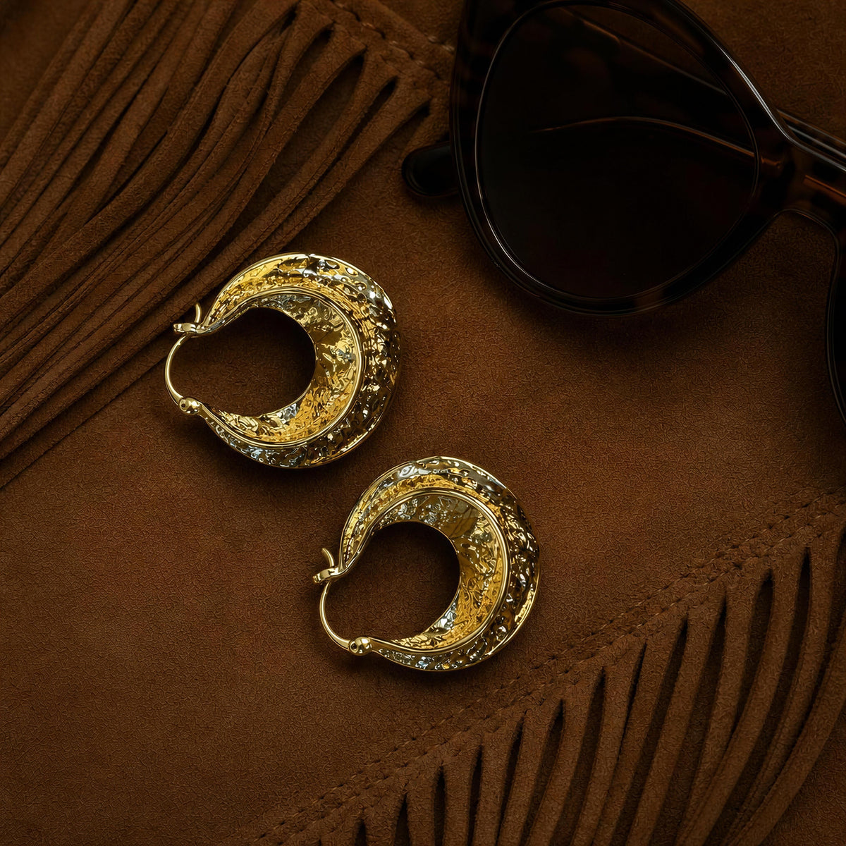 The Solea Earrings from  Chica Chica Jewelry , a pair of textured gold crescent-shaped hoop earrings displayed on a brown suede-like surface next to black sunglasses.
