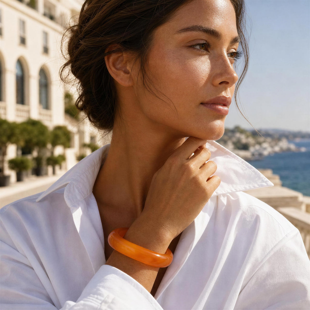 A woman in a white shirt wearing a translucent orange bangle bracelet, posing outdoors with a coastal city background.