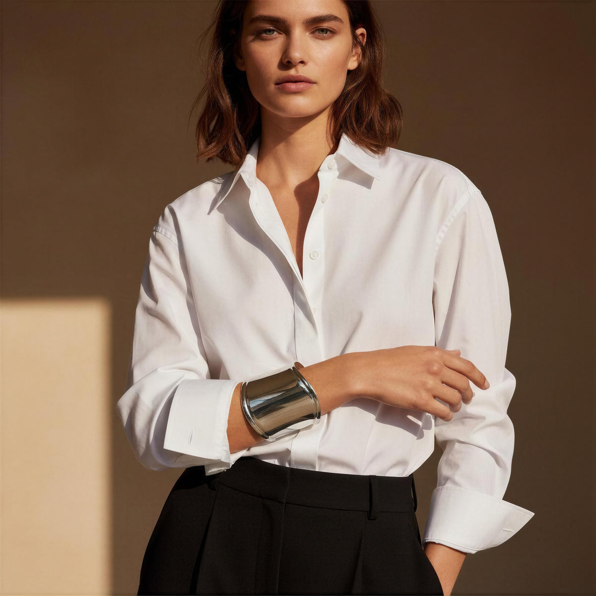 A woman wearing a crisp white long-sleeve button-down shirt tucked into black trousers, accessorized with the Roma Cuff from Chica Chica Jewelry, a chunky silver cuff bracelet, posed against a warm brown background.