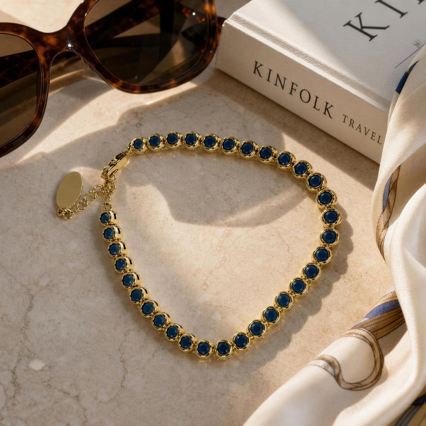 A gold tennis-style bracelet with turquoise gemstones, arranged on a light marble surface next to sunglasses, a book, and a white silk scarf.