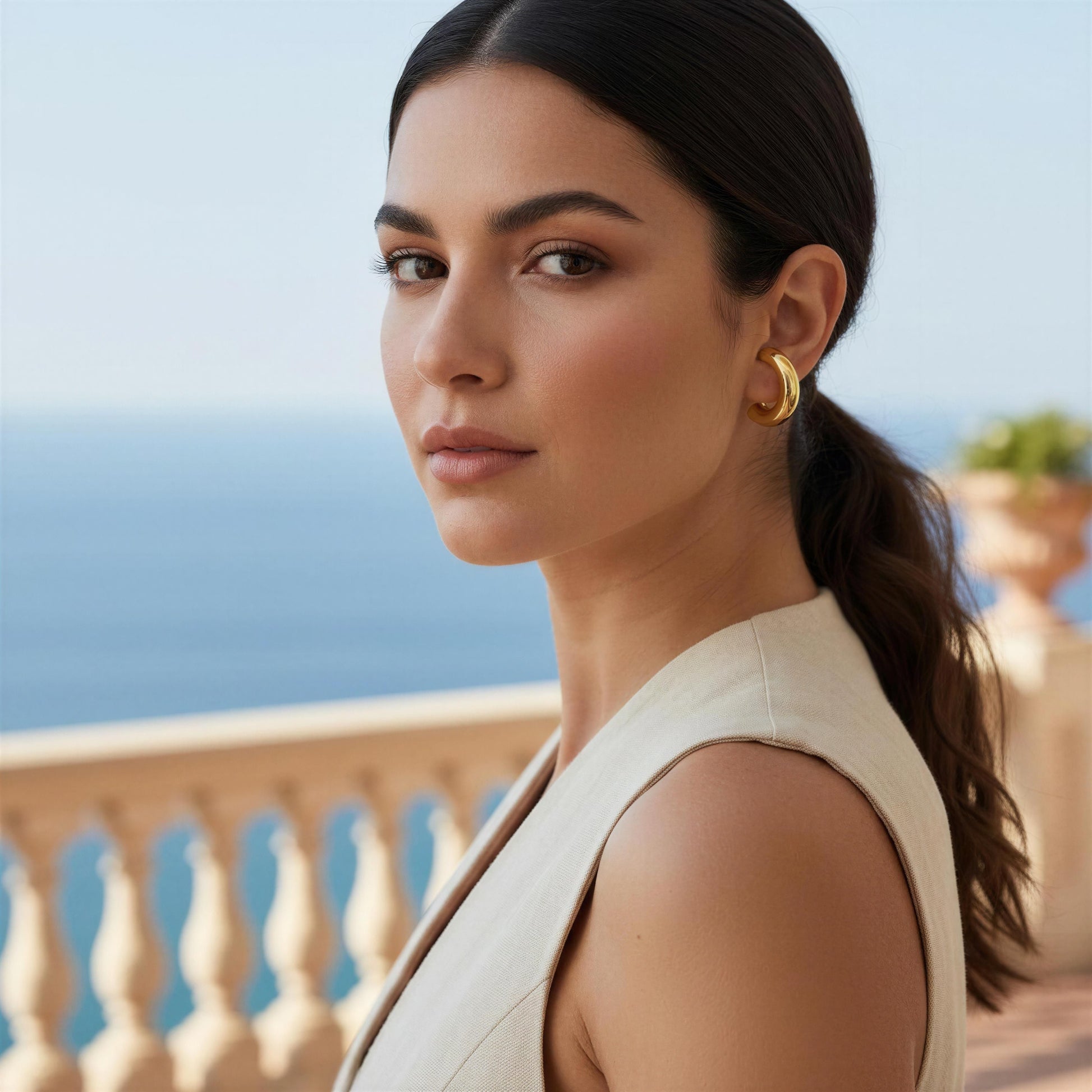 Woman with a ponytail and gold earring standing on a balcony with ocean view wearing the Stellar Arc Ear Cuff