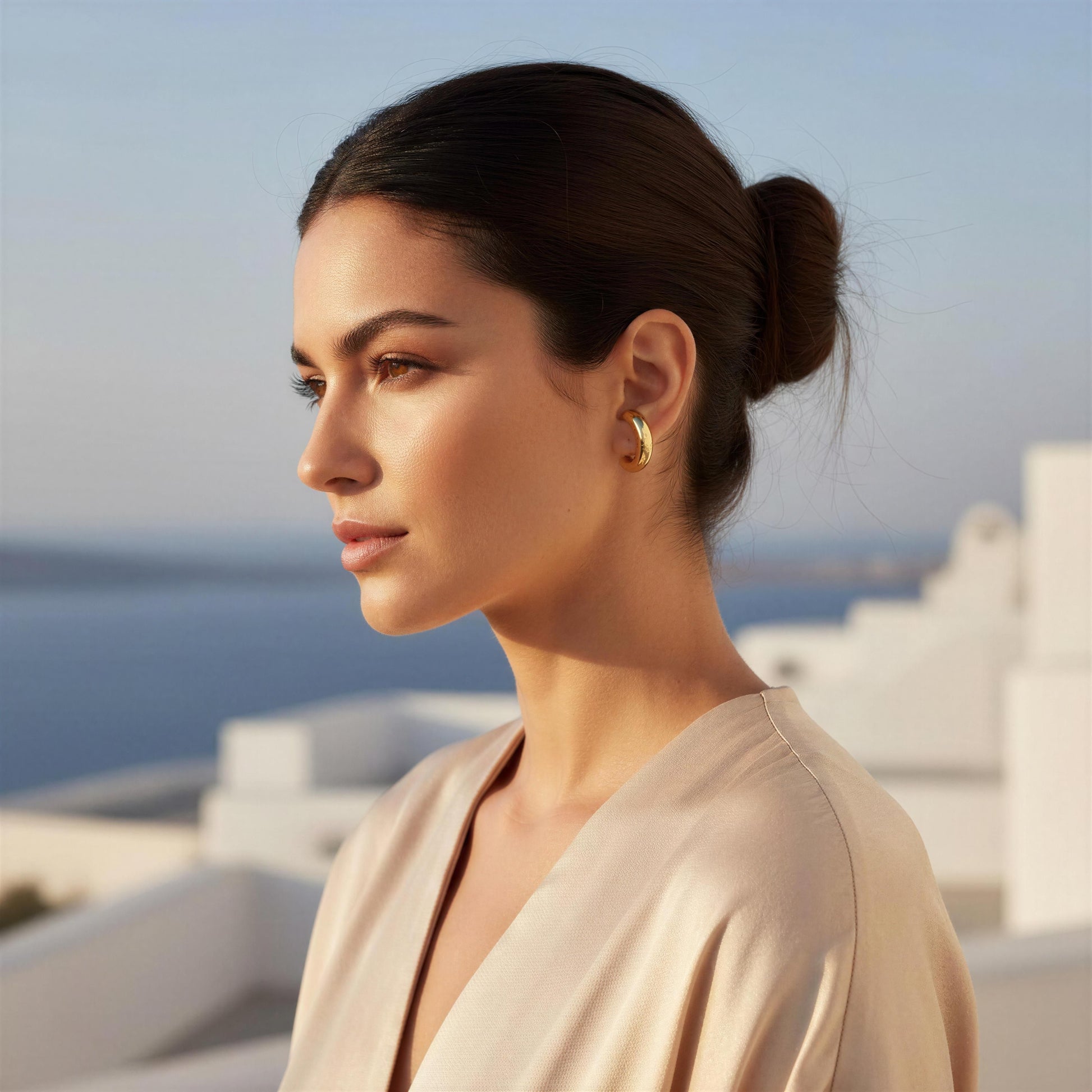 Woman with a bun hairstyle wearing gold earrings, standing against a scenic background.