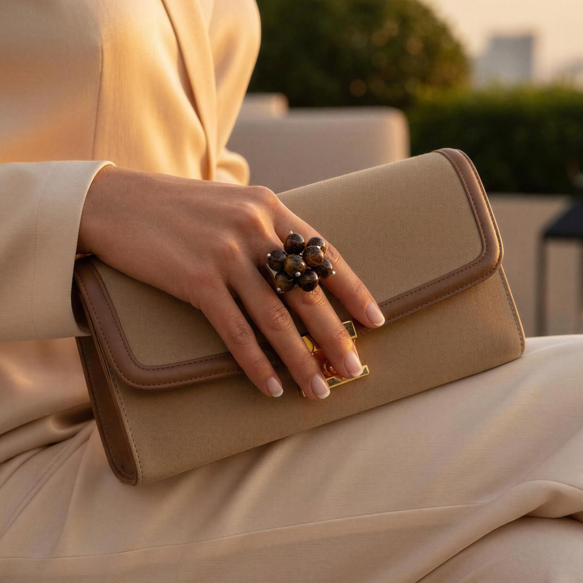 A woman in a beige satin outfit holds a beige leather clutch with a gold clasp on a rooftop terrace at sunset. She wears the Talia Ring from Chica Chica Jewelry.