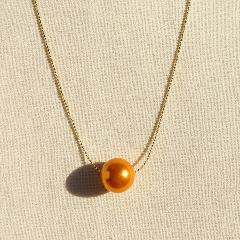 The Tangerine Petite Sphere Necklace_ from Chica Chica Jewelry with a single orange bead on a beige background