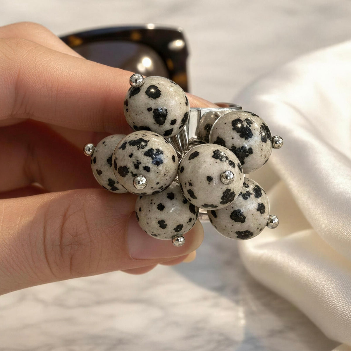 A hand holding the Dalma Ring  from Chica Chica Jewelry -  a cluster of round white beads with black spots, resembling dalmatian-style stones, with small silver metal accents. The background includes a white satin fabric and a marble surface.