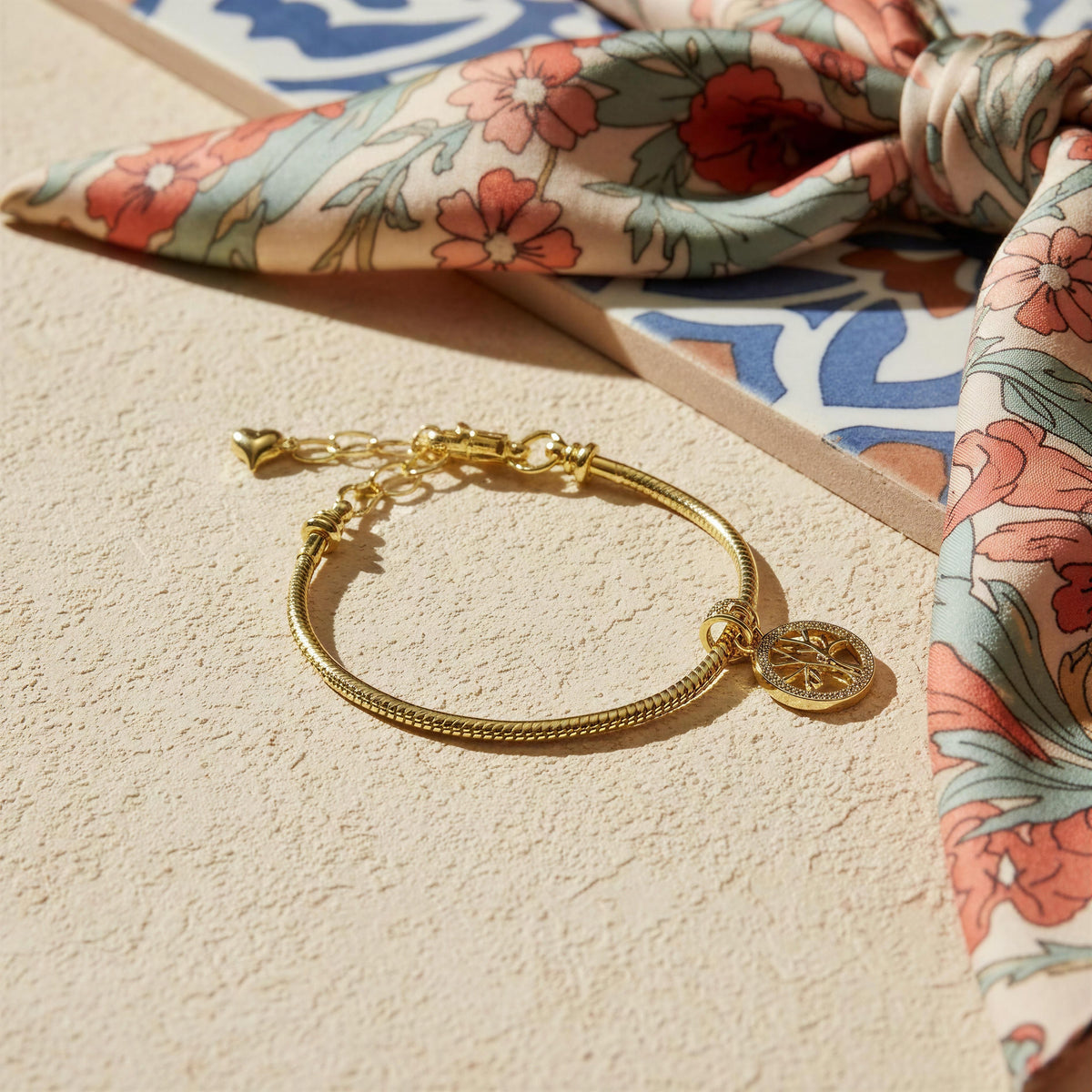 A gold-toned snake chain bracelet with three of life charm, styled on a textured beige surface next to a floral silk scarf.
