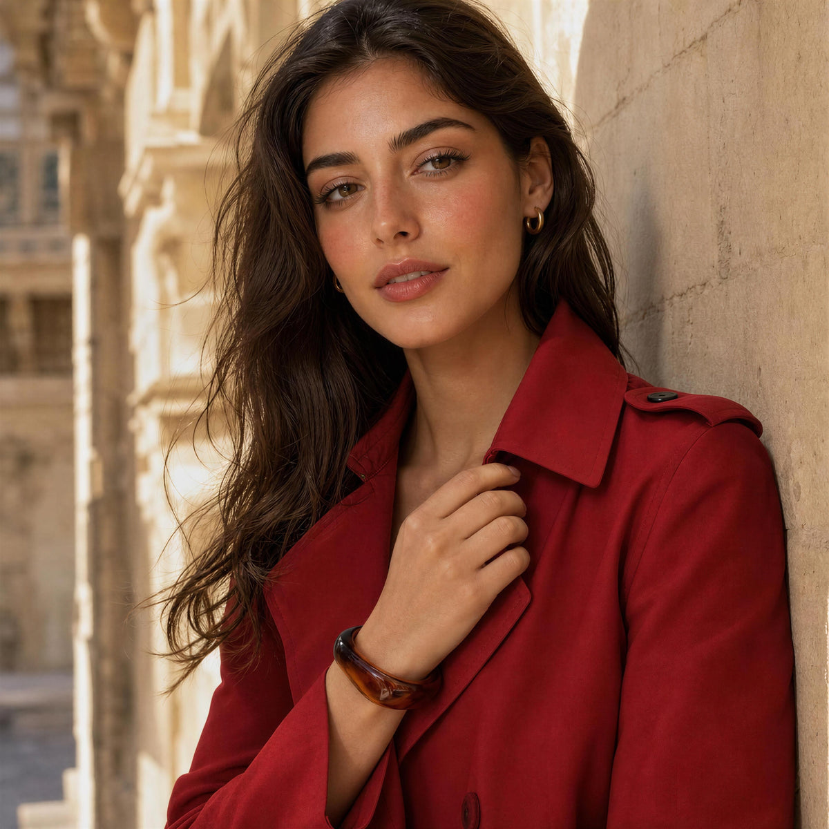 A woman with long dark brown hair leans against a beige stone wall, wearing a vibrant red trench coat and a brown tortoiseshell bracelet.