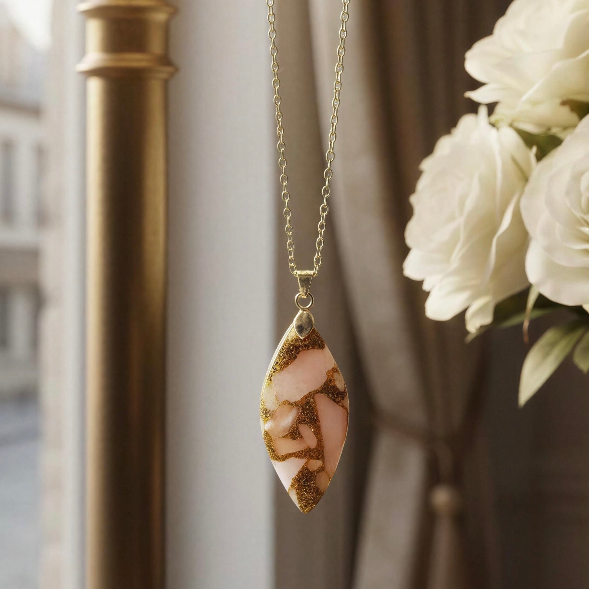 A gold chain necklace with a marquise-shaped pendant featuring a pink and white marbled pattern with gold veins, displayed against a soft-focus background with white roses and a gold column.