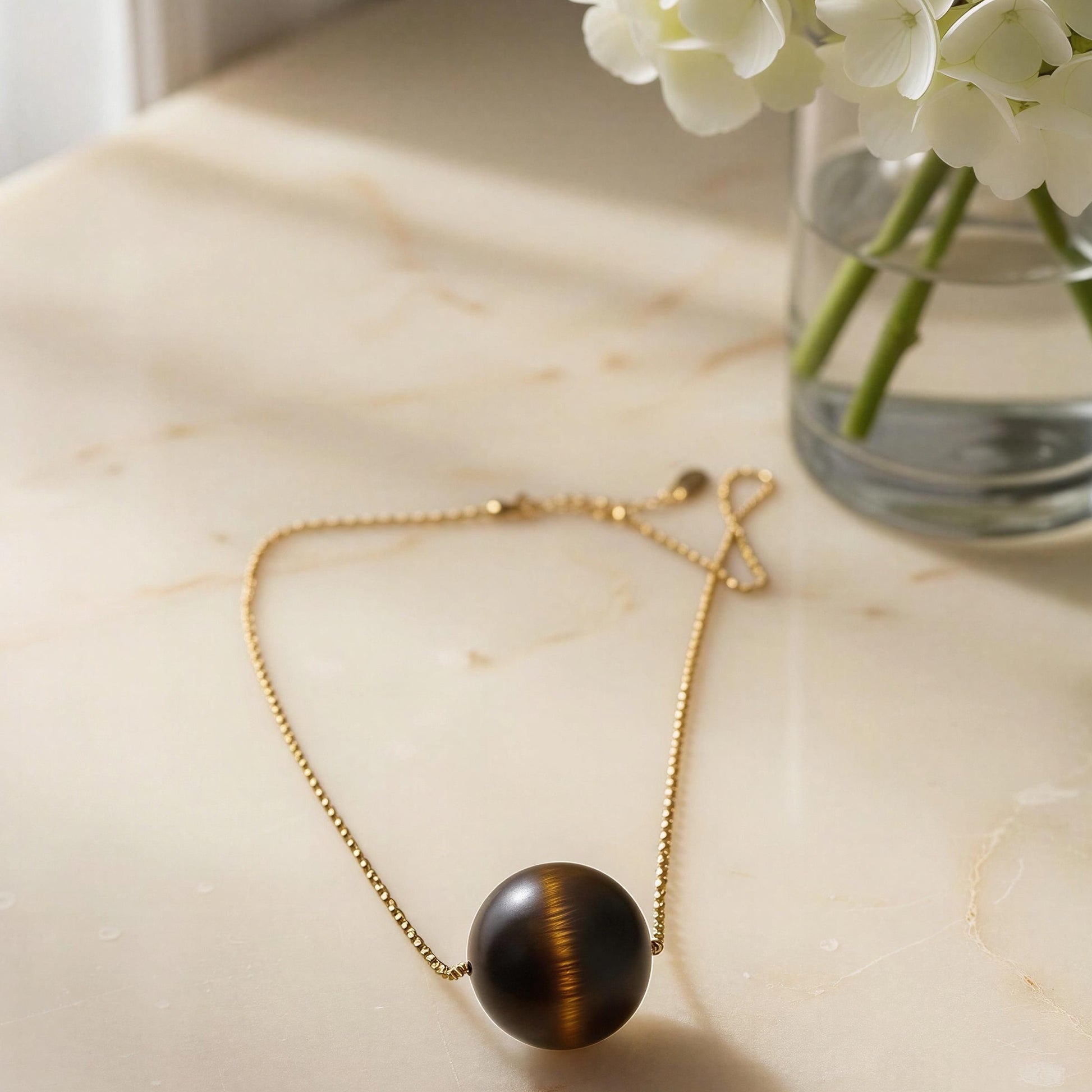 The Terra Sol Sphere Necklace  on a marble surface next to a vase of white flowers.