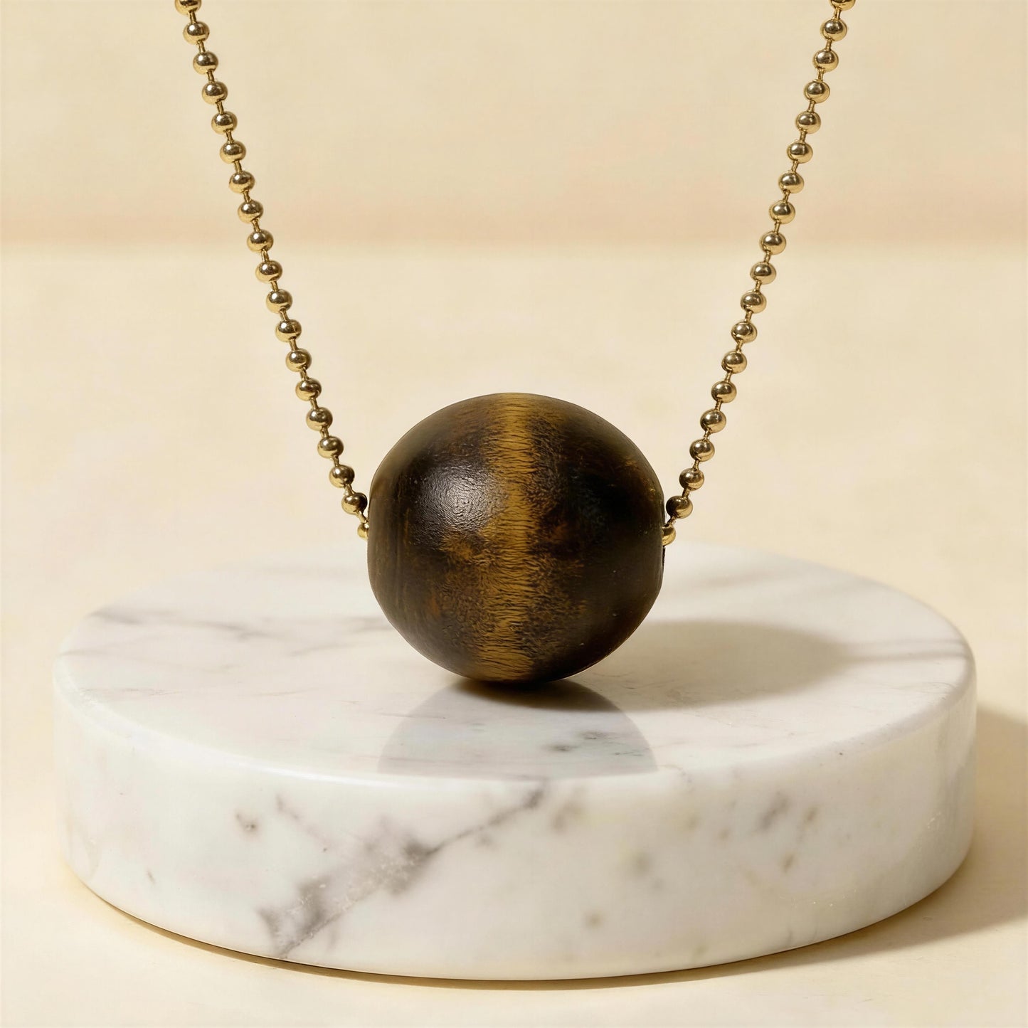 Wooden sphere pendant on a gold ball chain on a marble stand with a beige background