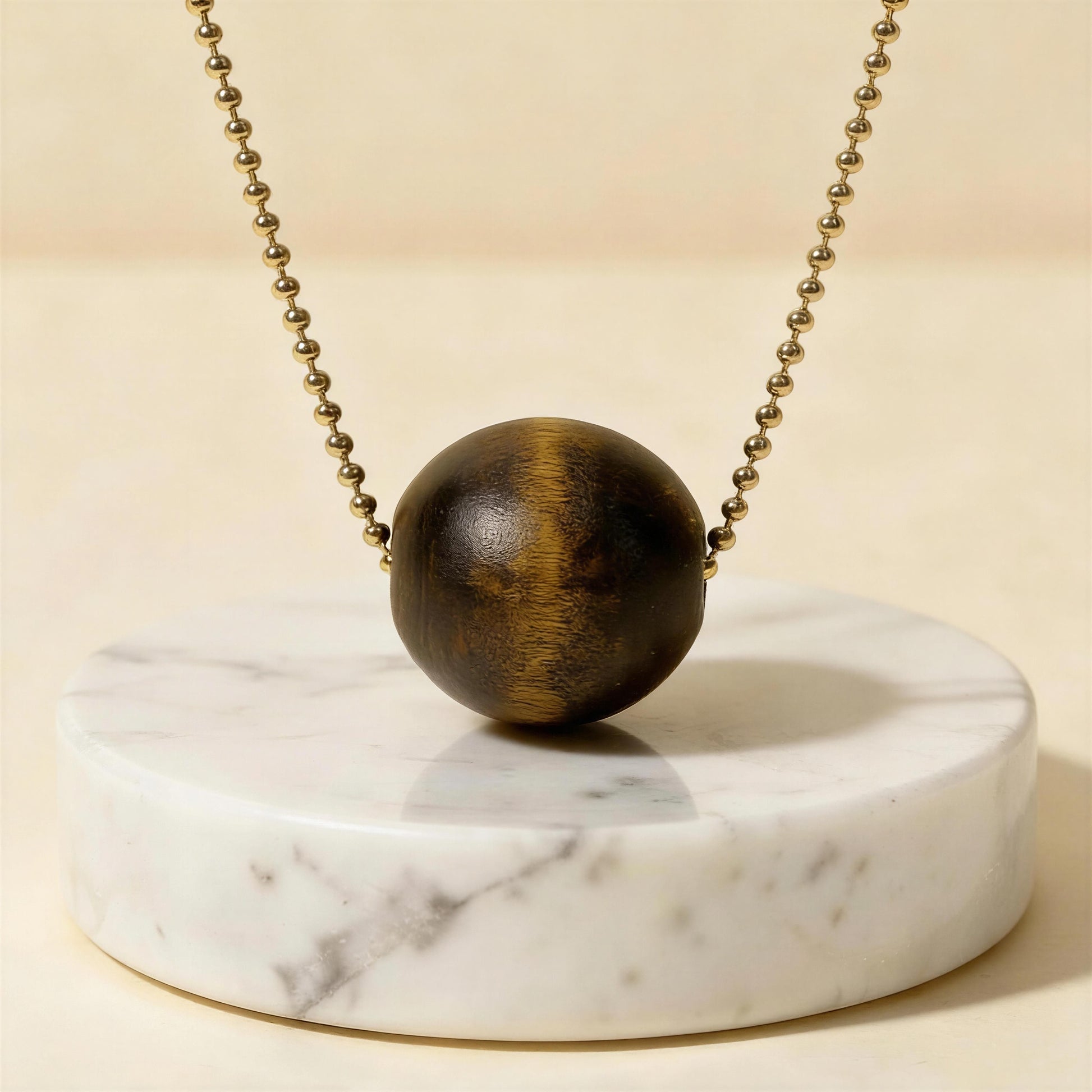 Wooden sphere pendant on a gold ball chain on a marble stand with a beige background