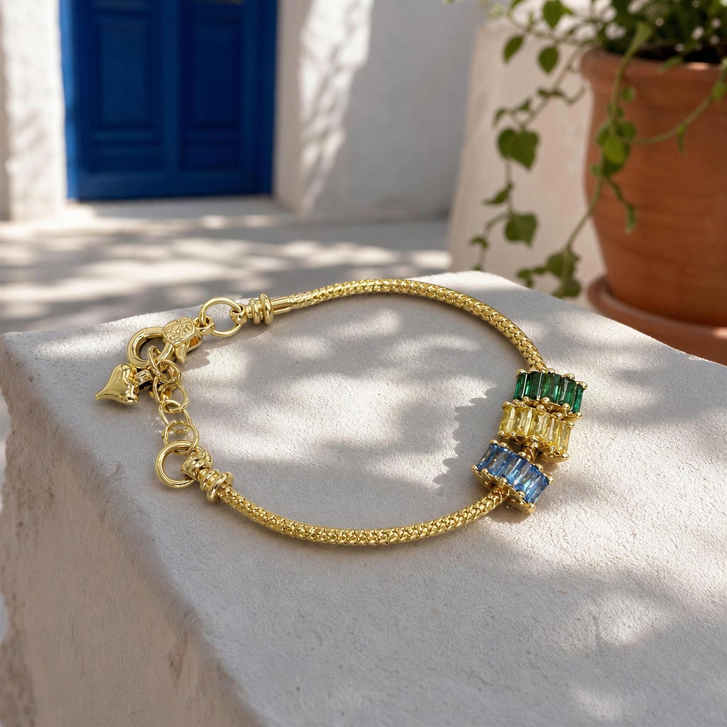 A gold chain bracelet with three stacked rectangular bead charms in green, yellow, and blue, resting on a white stone pedestal with a Mediterranean-style background.