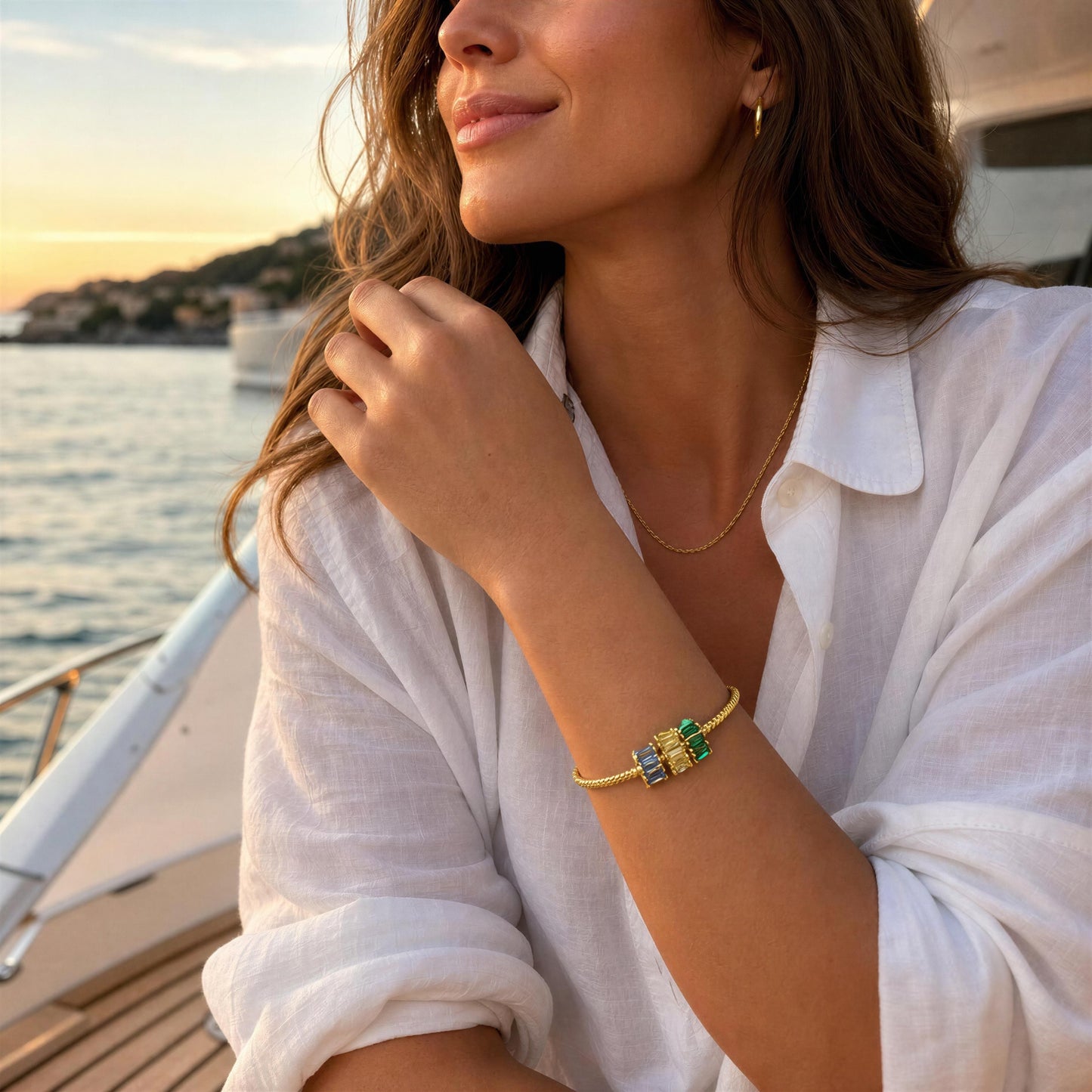 A woman in a white linen shirt on a boat at sunset, wearing a delicate a gold bracelet with green, yellow and blue crystal accents.