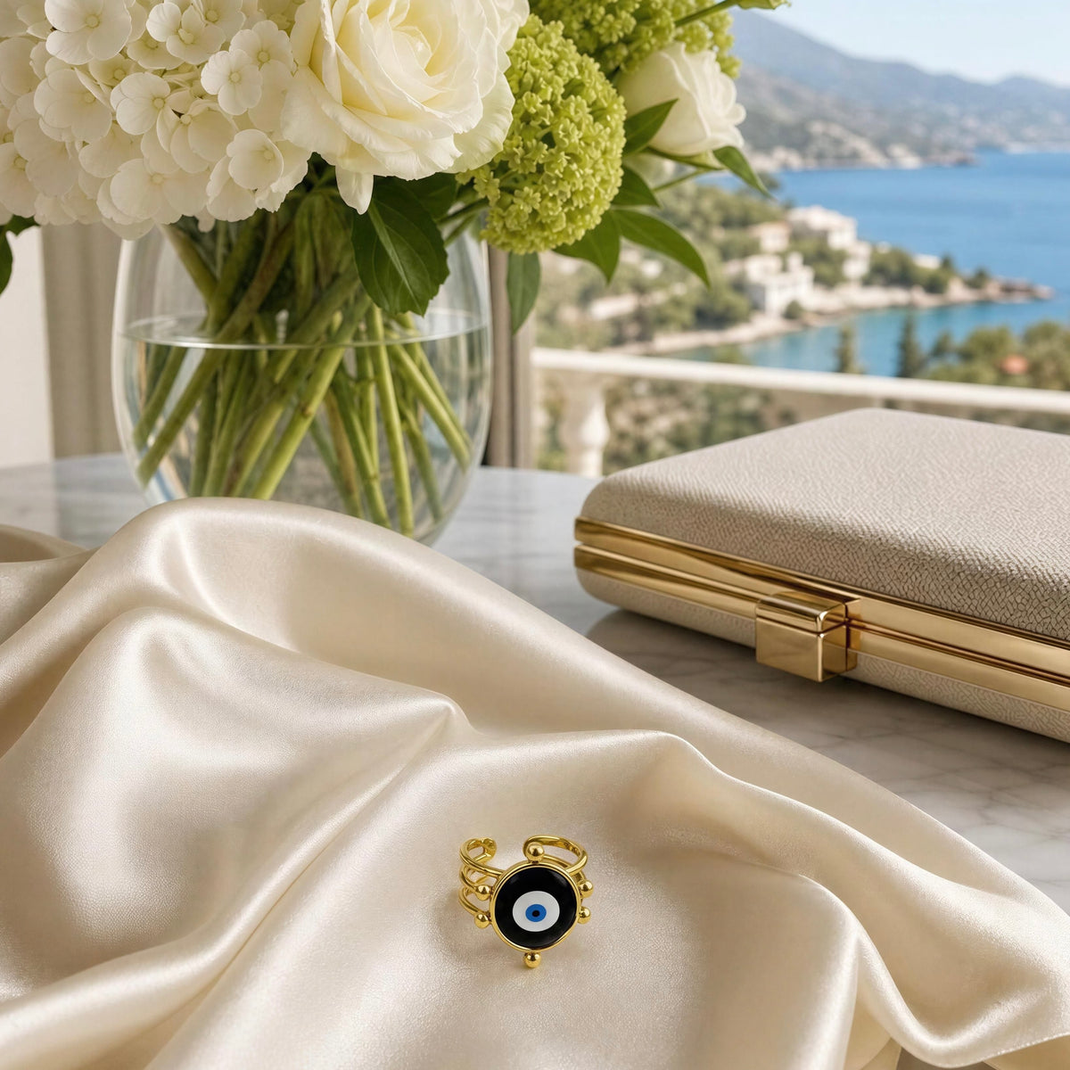 A gold ring with a black evil eye pendant and blue center, styled on draped champagne satin next to a beige clutch with gold trim, with a vase of white and green flowers and a coastal balcony view in the background.
