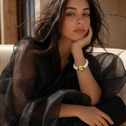 A woman in a black sheer blouse rests her head on her hand, wearing a white beaded bracelet with a yellow smiley face charm.
