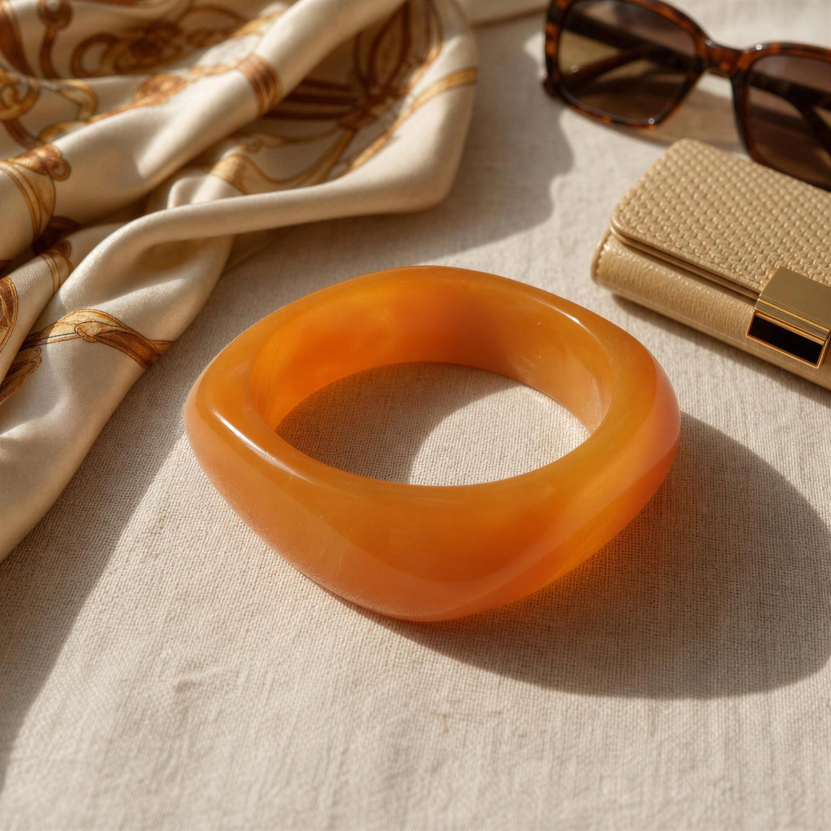 A chunky, thick orange translucent resin bangle bracelet displayed on a textured beige fabric surface, styled with a patterned cream scarf, tortoiseshell sunglasses, and a beige clutch bag.