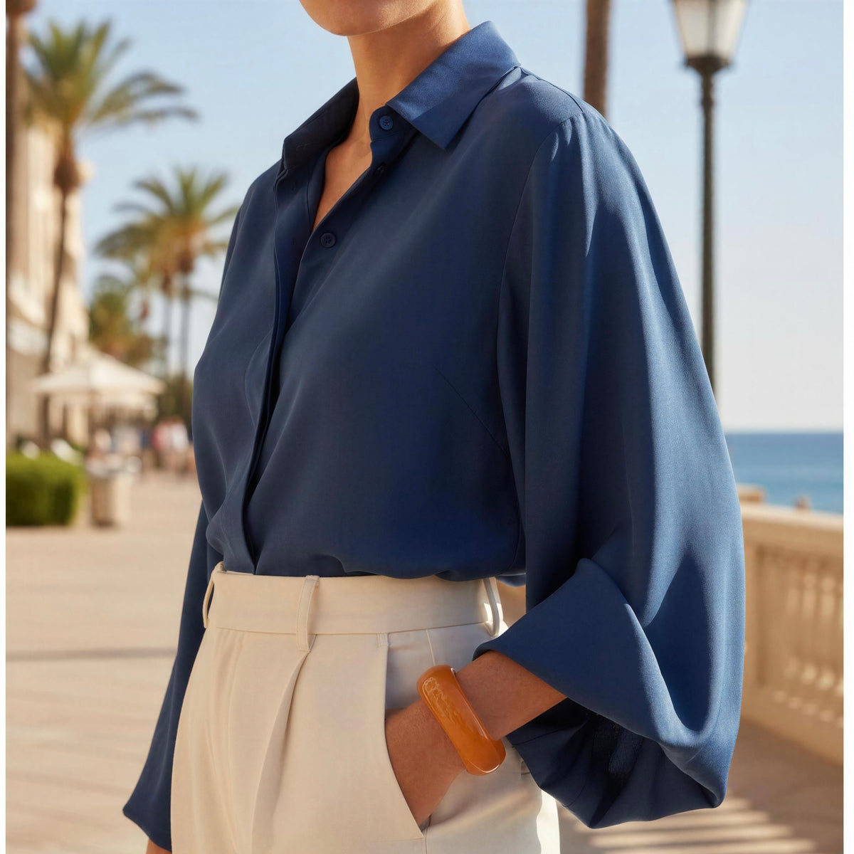 A woman wearing a navy blue long-sleeve button-up blouse tucked into cream high-waisted trousers, standing on a sunny seaside promenade.
