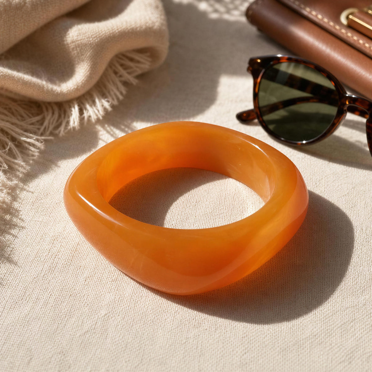A translucent orange bangle bracelet with a thick, rounded square shape, styled on a beige textured fabric alongside sunglasses, a brown leather accessory, and a cream scarf.