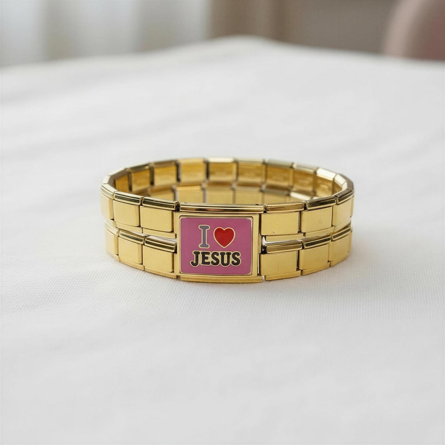 A gold-tone metal link bracelet with a rectangular center plate that reads 'I love JESUS' on a pink background, photographed on a white fabric surface.