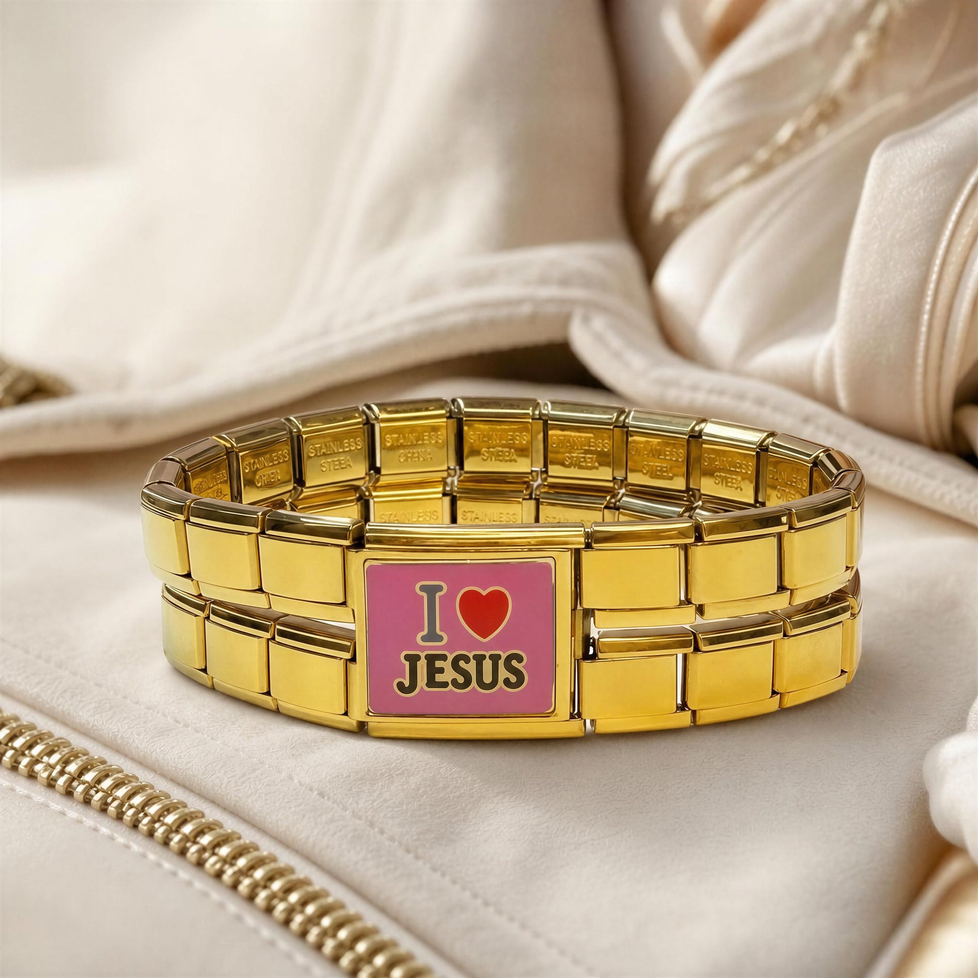 A gold-tone metal link bracelet with a rectangular pink center plate that reads 'I love JESUS' in gold lettering, displayed on a soft cream fabric surface.