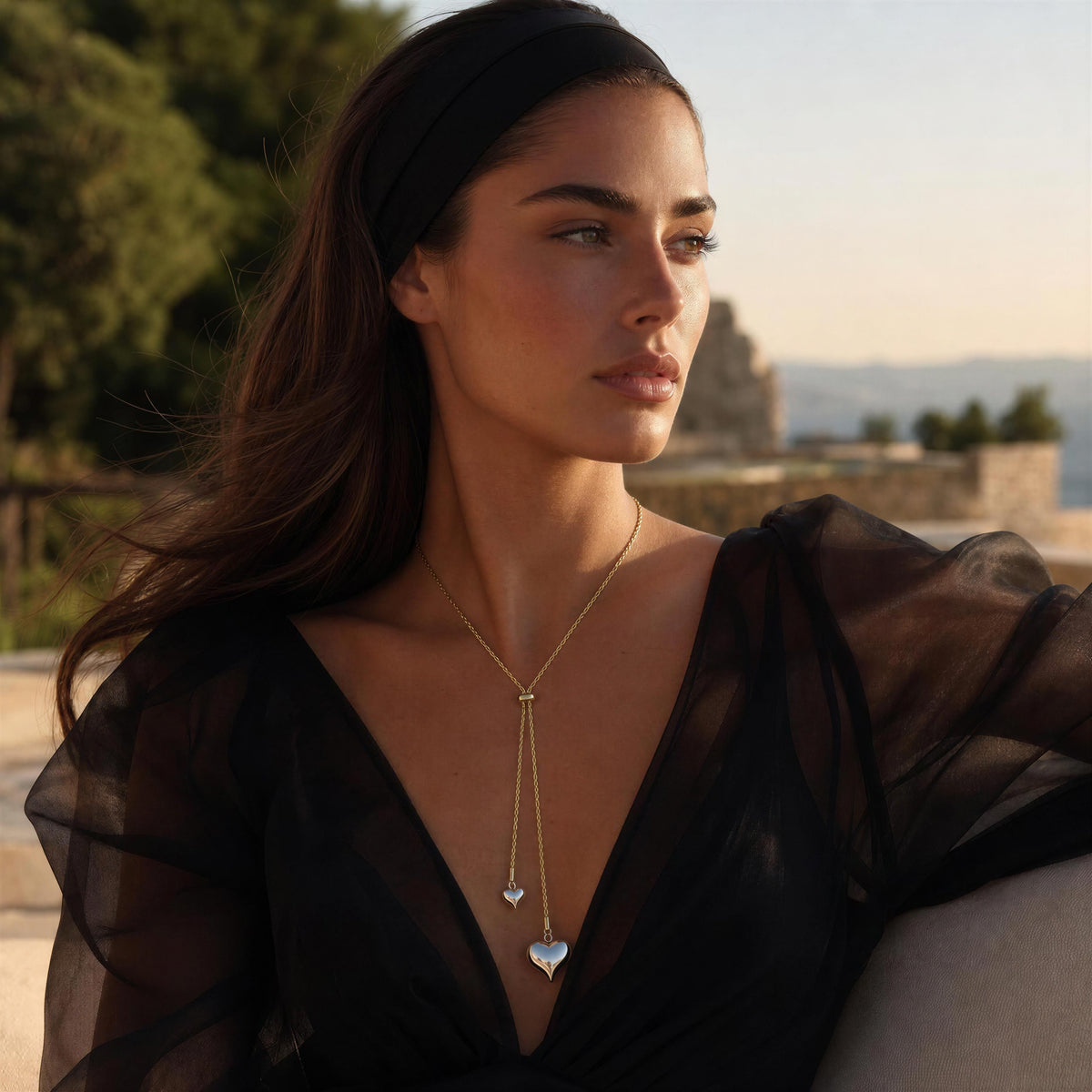 A woman wearing the Amanti Lariat Necklace, a delicate gold Y-necklace with two silver heart pendants, styled with a black sheer top and headband in an outdoor setting.