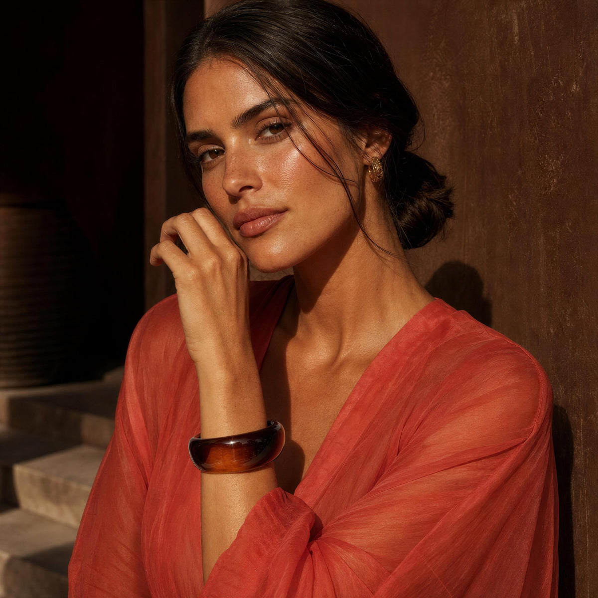 A woman in a sheer coral-orange blouse leans against a dark wooden wall, wearing the Ambra Bangle from Chica Chica Jewelry, a chunky amber-brown tortoiseshell bangle on her wrist.