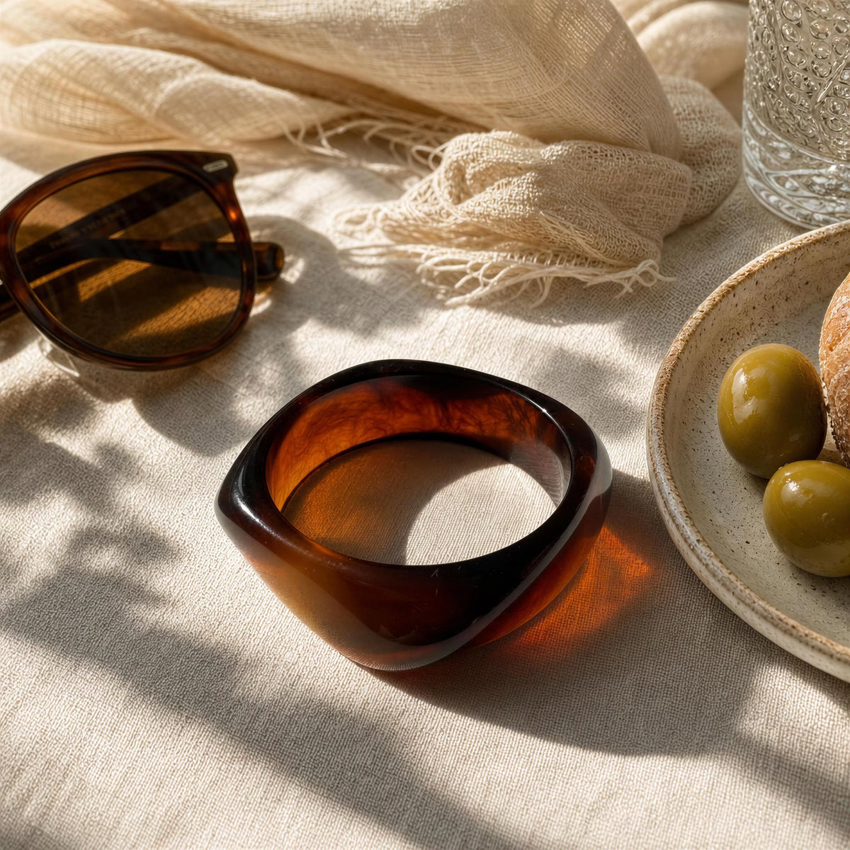 A thick, chunky tortoiseshell bangle bracelet in a warm amber-brown color, styled on a beige linen tablecloth alongside tortoiseshell sunglasses, a cream scarf, a glass of water, and a plate with olives.