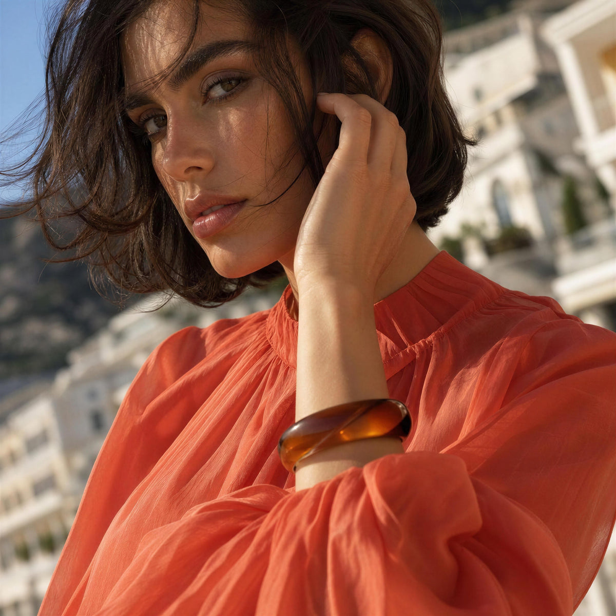 A woman with short dark hair wearing a sheer coral-orange blouse and the Ambra Bangle from Chica Chica Jewelry,  a translucent amber-brown bangle bracelet, photographed outdoors with a blurred coastal building background.