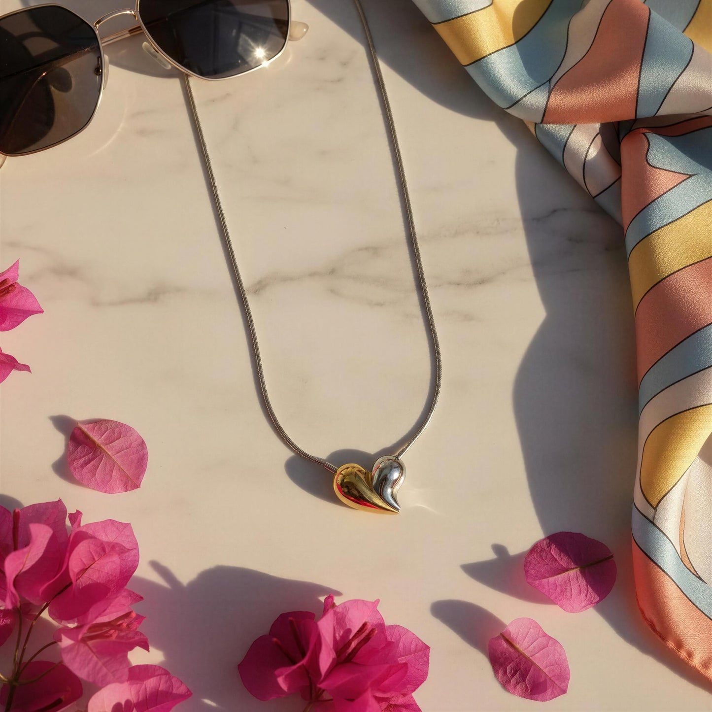 Necklace with heart-shaped pendant, sunglasses, and colorful scarf on a marble surface with pink flowers.