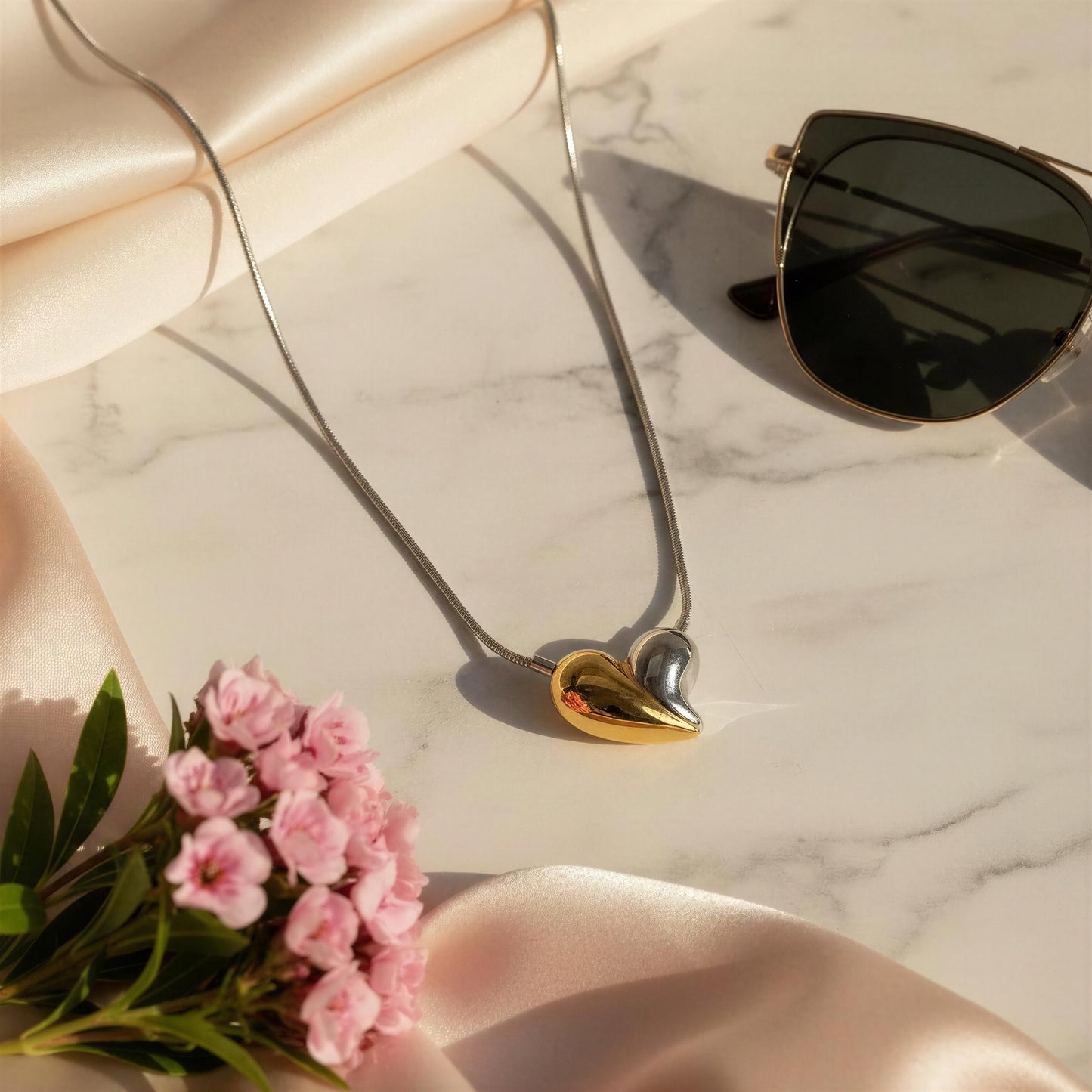The  Amor Eterno Necklace, a heart-shaped pendant, sunglasses, and pink flowers on a marble surface