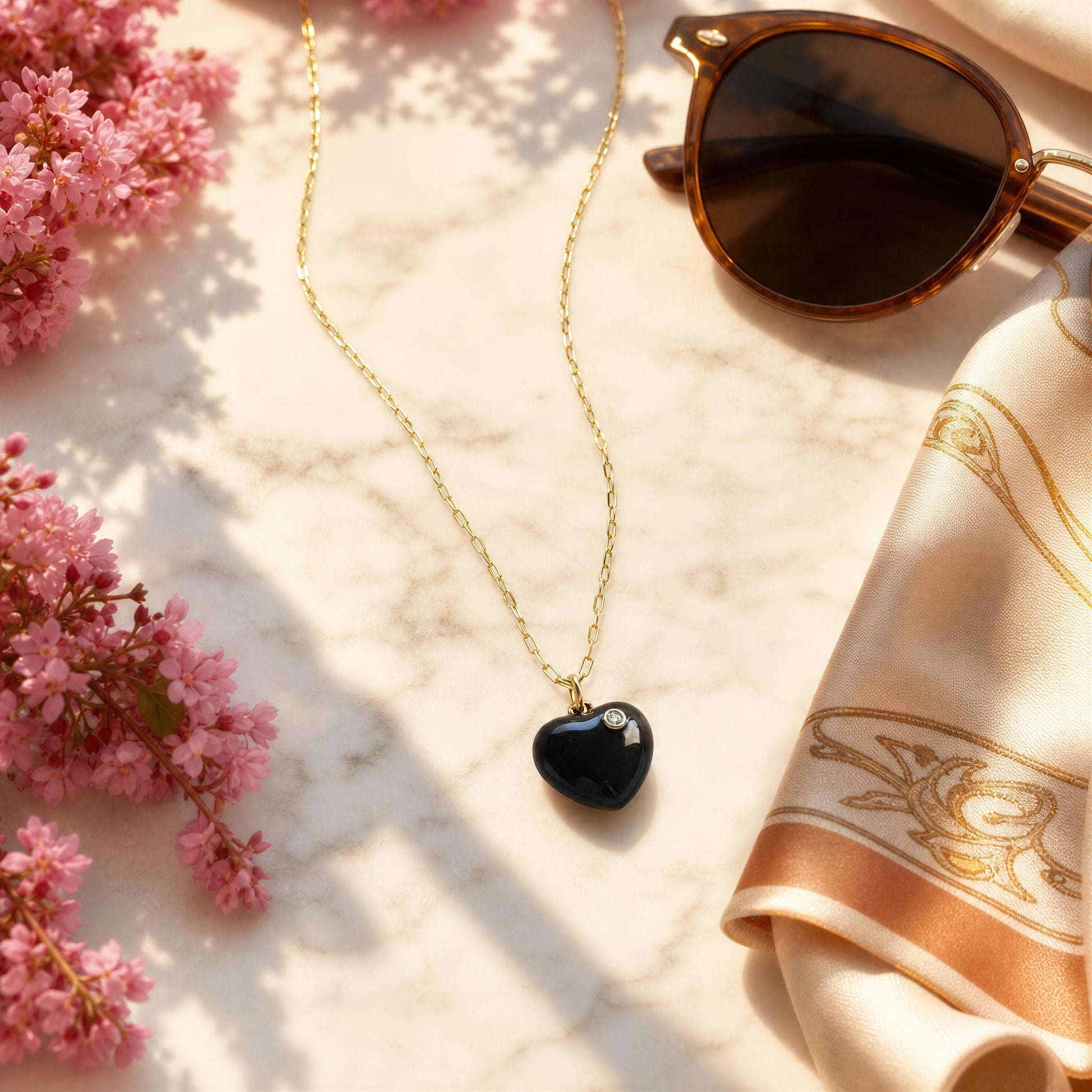 Necklace with a heart-shaped pendant, sunglasses, and a scarf on a marble surface with pink flowers.