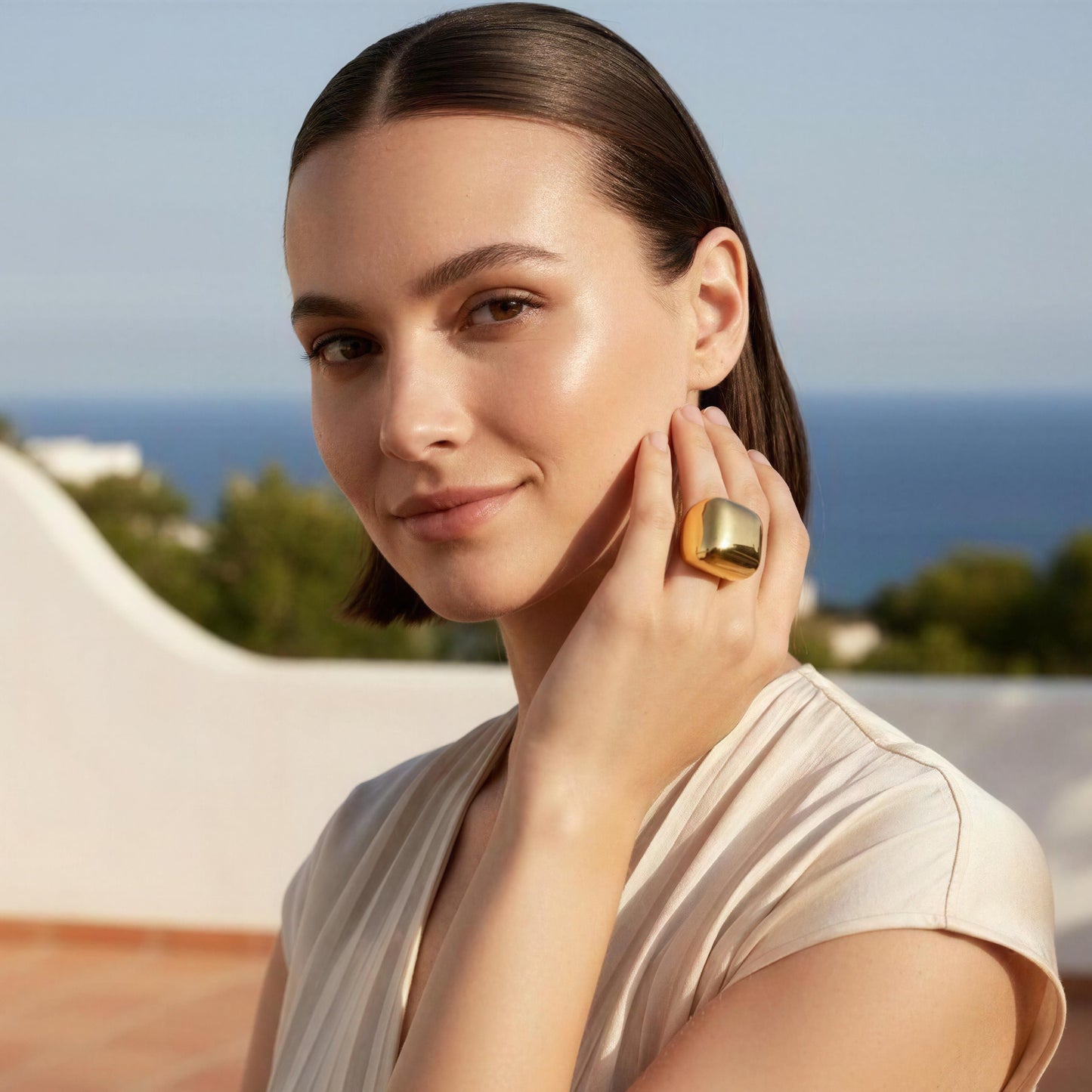 A woman with dark hair pulled back and the Aurelia Ring from Chica Chica Jewelry  on her finger, posing outdoors with a white wall and blue sea in the background.