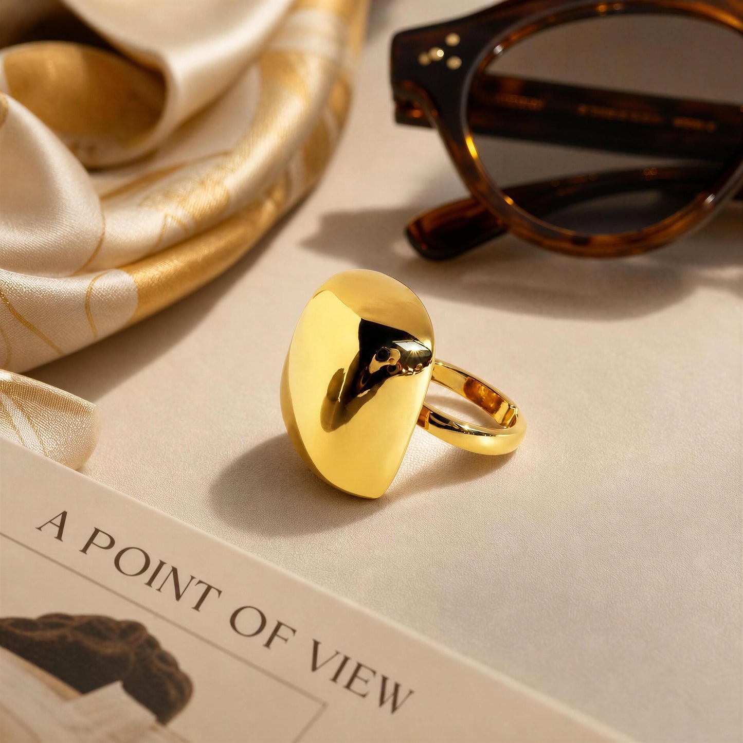 The Aurelia Ring from Chica Chica Jewelry styled on a beige surface with a cream silk scarf, tortoiseshell sunglasses, and a book page.