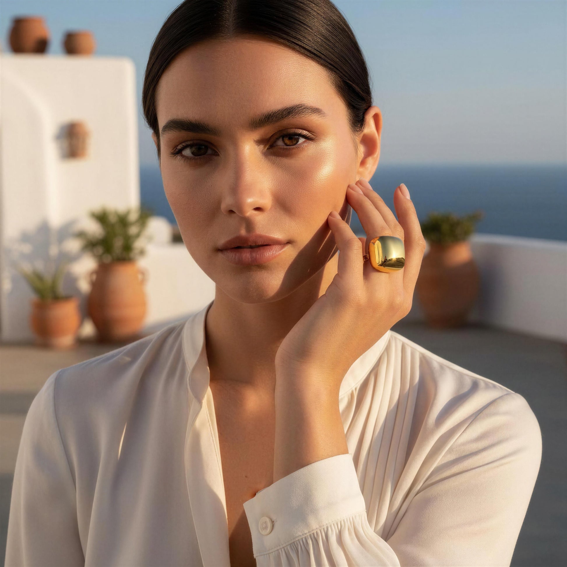 A woman in a white blouse wearing the Aurelia Ring from Chica Chica Jewelry , posing outdoors with a white wall and sea in the background.
